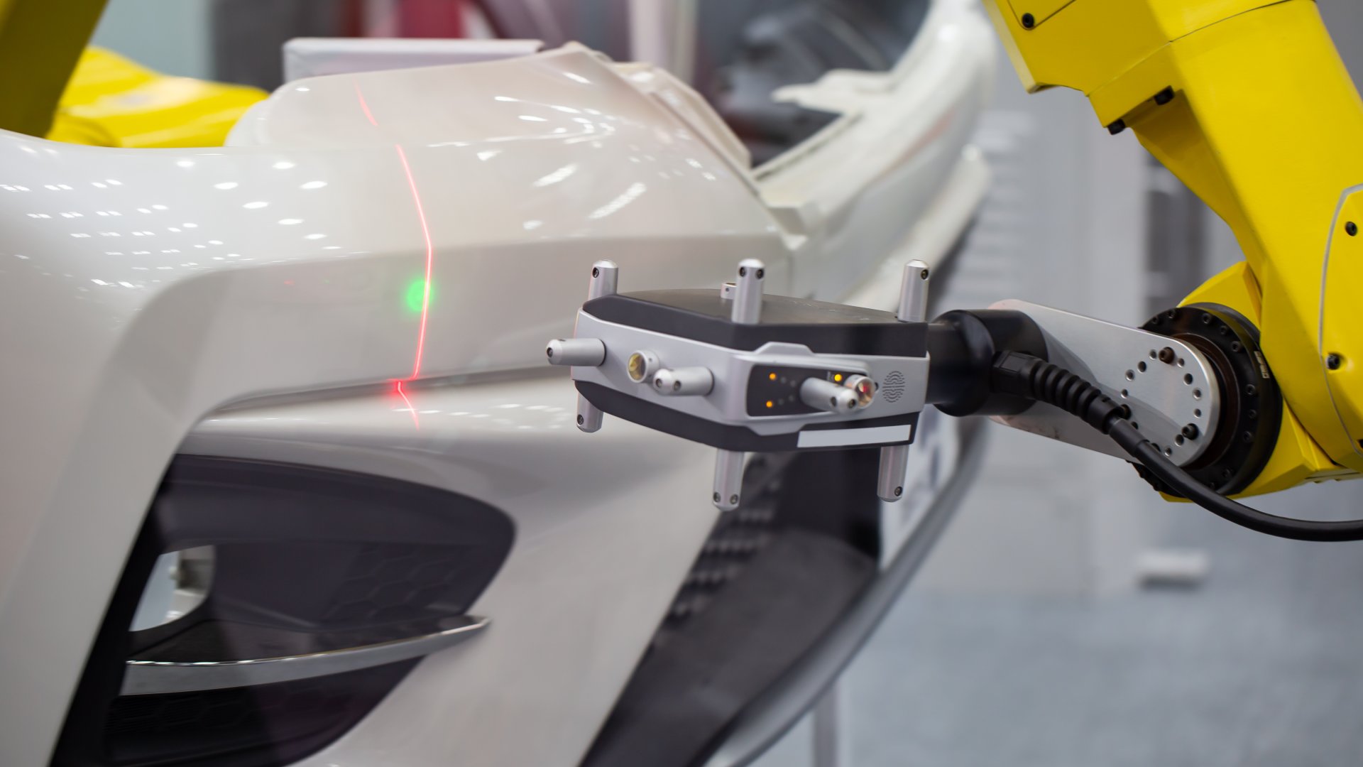 A yellow robotic arm uses a laser tracking tool to measure the dimensions of a white bumper to a car.