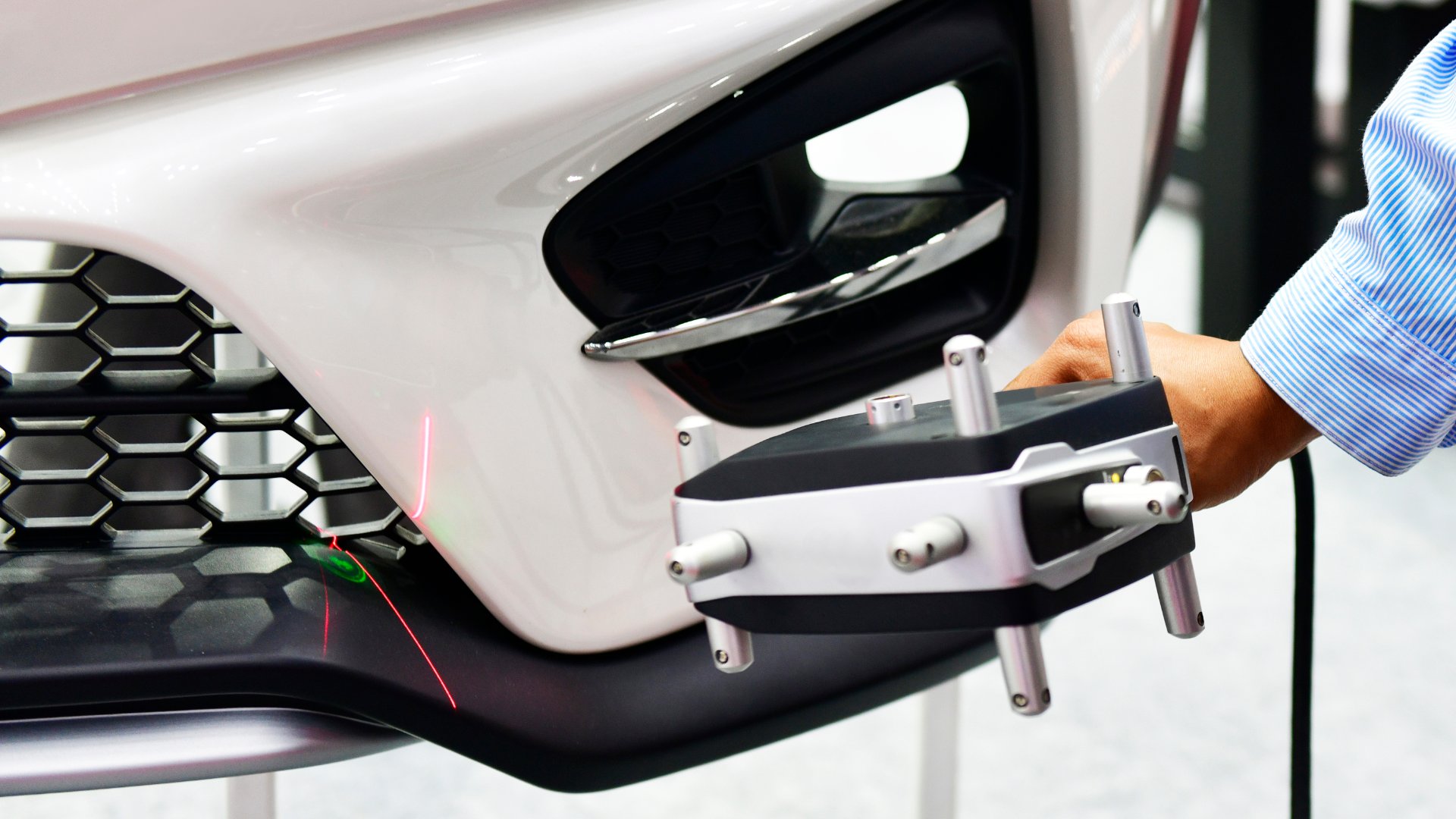 A close-up of the front white bumper of a vehicle being manufactured. Someone holds a laser scanner up to the bumper.