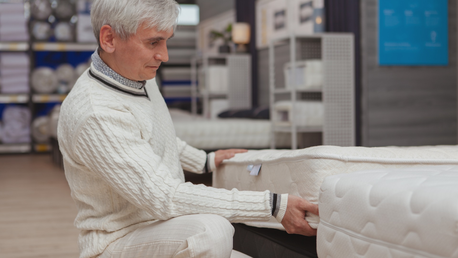 A senior man shops for a mattress at the store. He kneels next to a mattress option and inspects the thickness.