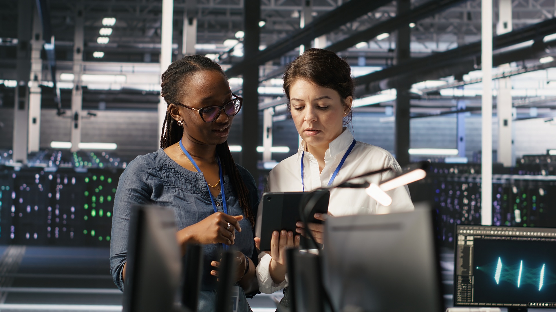 Two co-workers standing side-by-side and working together to review infomation on a tablet in a large server room.