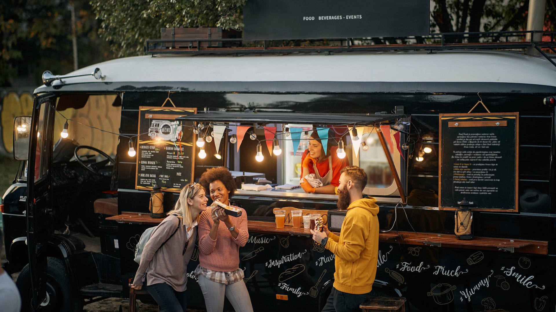 A group of three individuals share food in front of a stylish, modern-looking food truck. A worker smiles at them from inside.