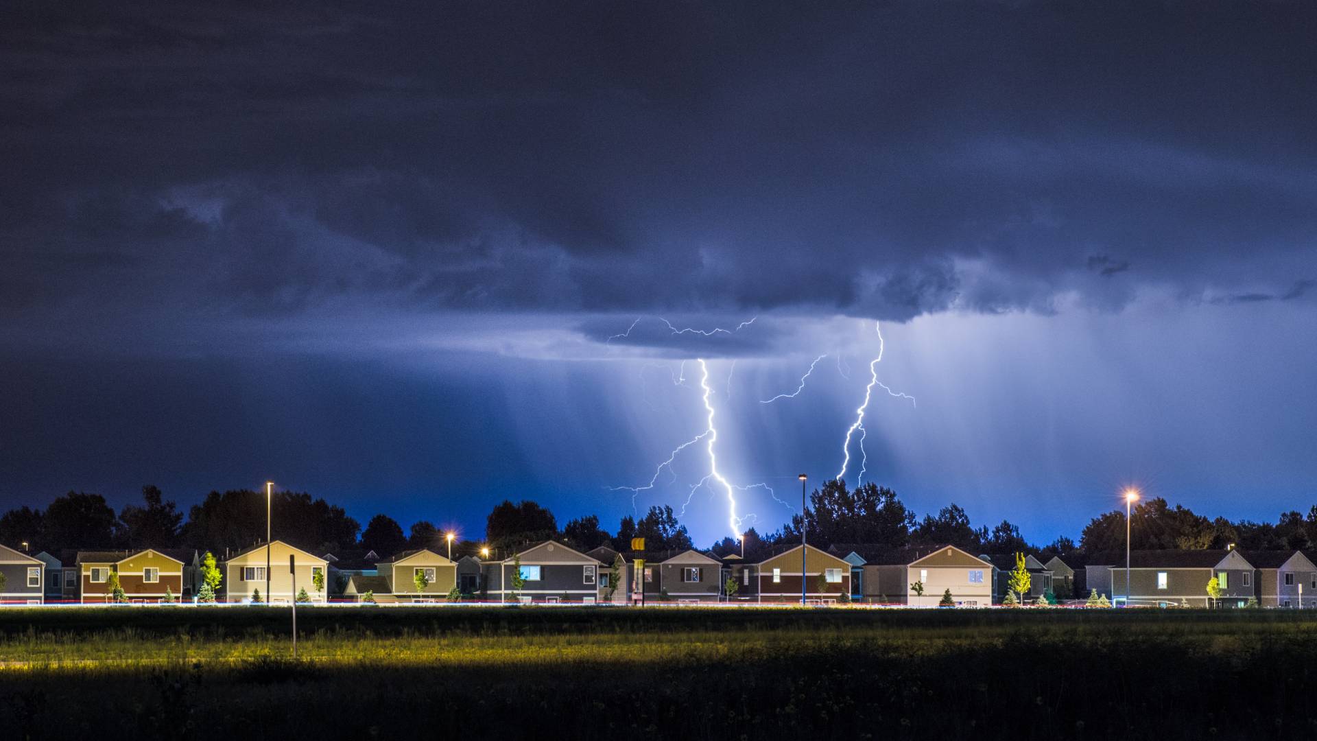 A neighborhood at night is lit up by lightning strikes coming from the storm clouds above them. It's also raining.