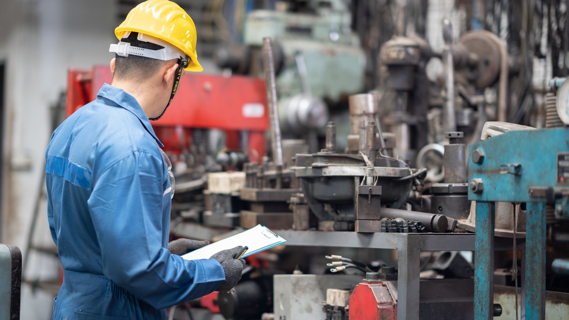 A manufacturing worker wearing a hard hat stands in front of a machine in a factory. He holds a clipboard.