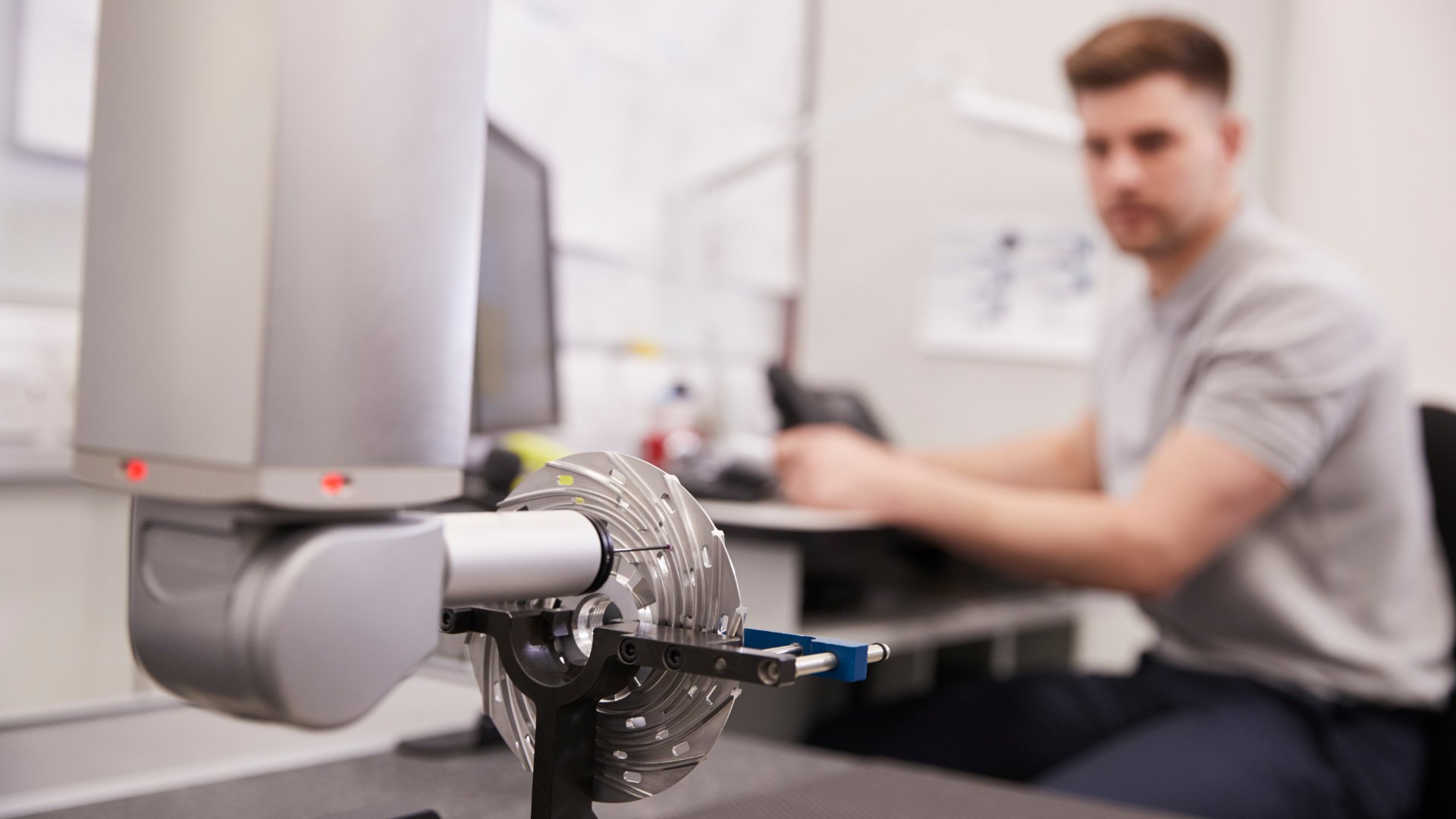 A CMM performing a measurement routine on a part. A man sits at a computer desk, blurred in the background.