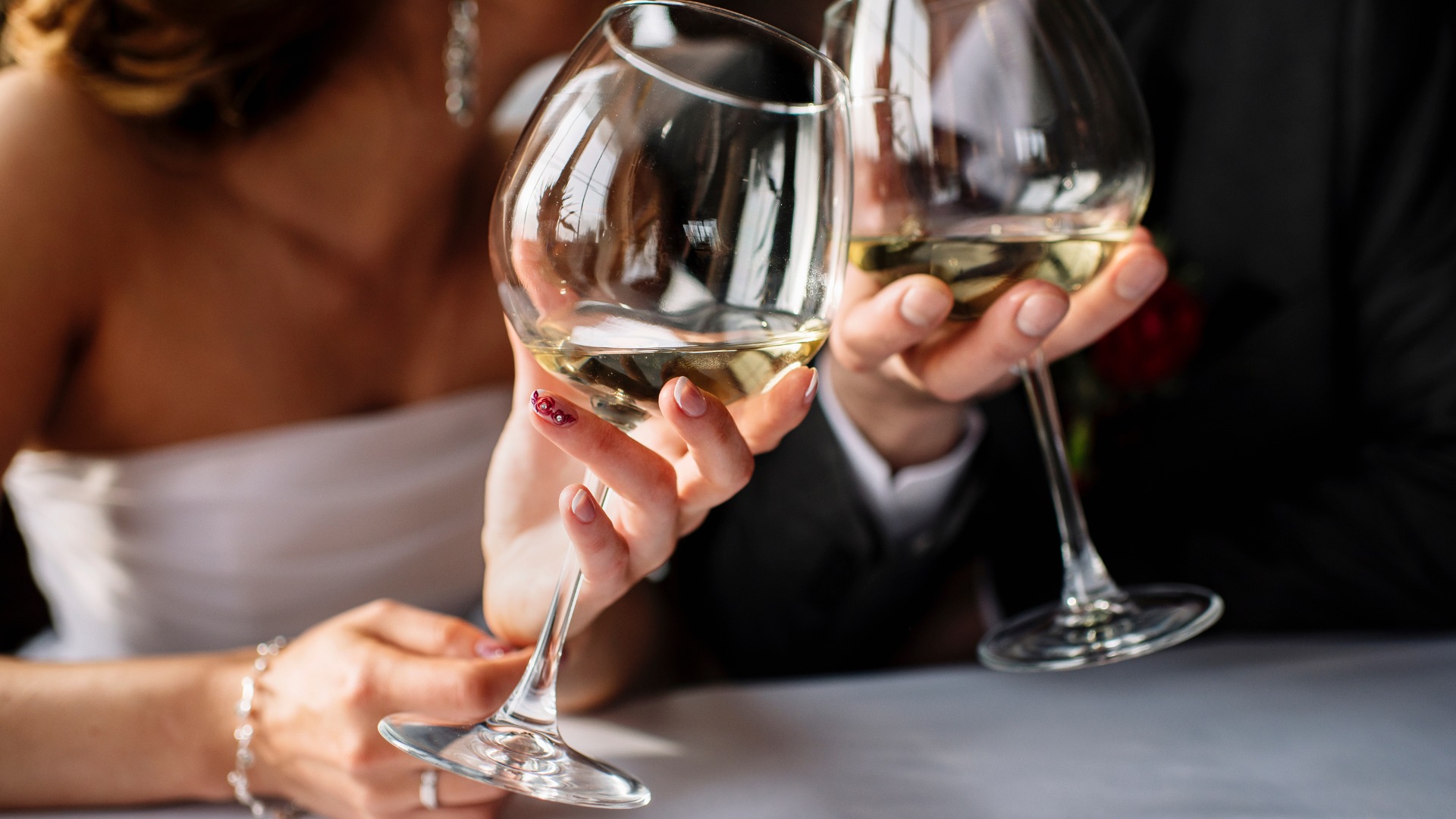 A bride and groom sitting at a table while toasting wine glasses. The glasses have white wine inside them.