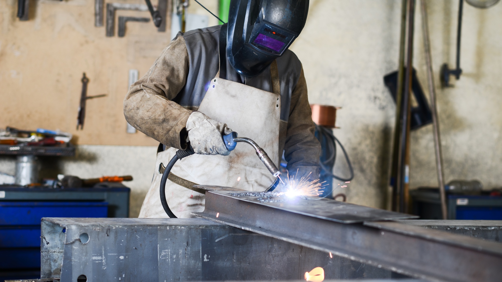 A man wearing protective gear and a helmet welds a large metal frame with a MIG welding gun on a table in a workshop.