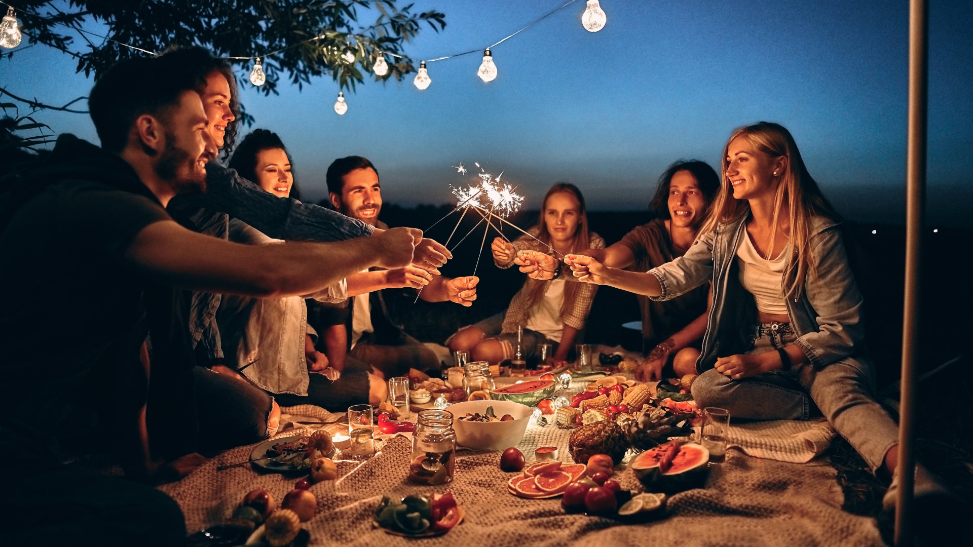  Seven friends sit around a small blanket full of food outside. They all hold sparklers above the middle of the blanket.
