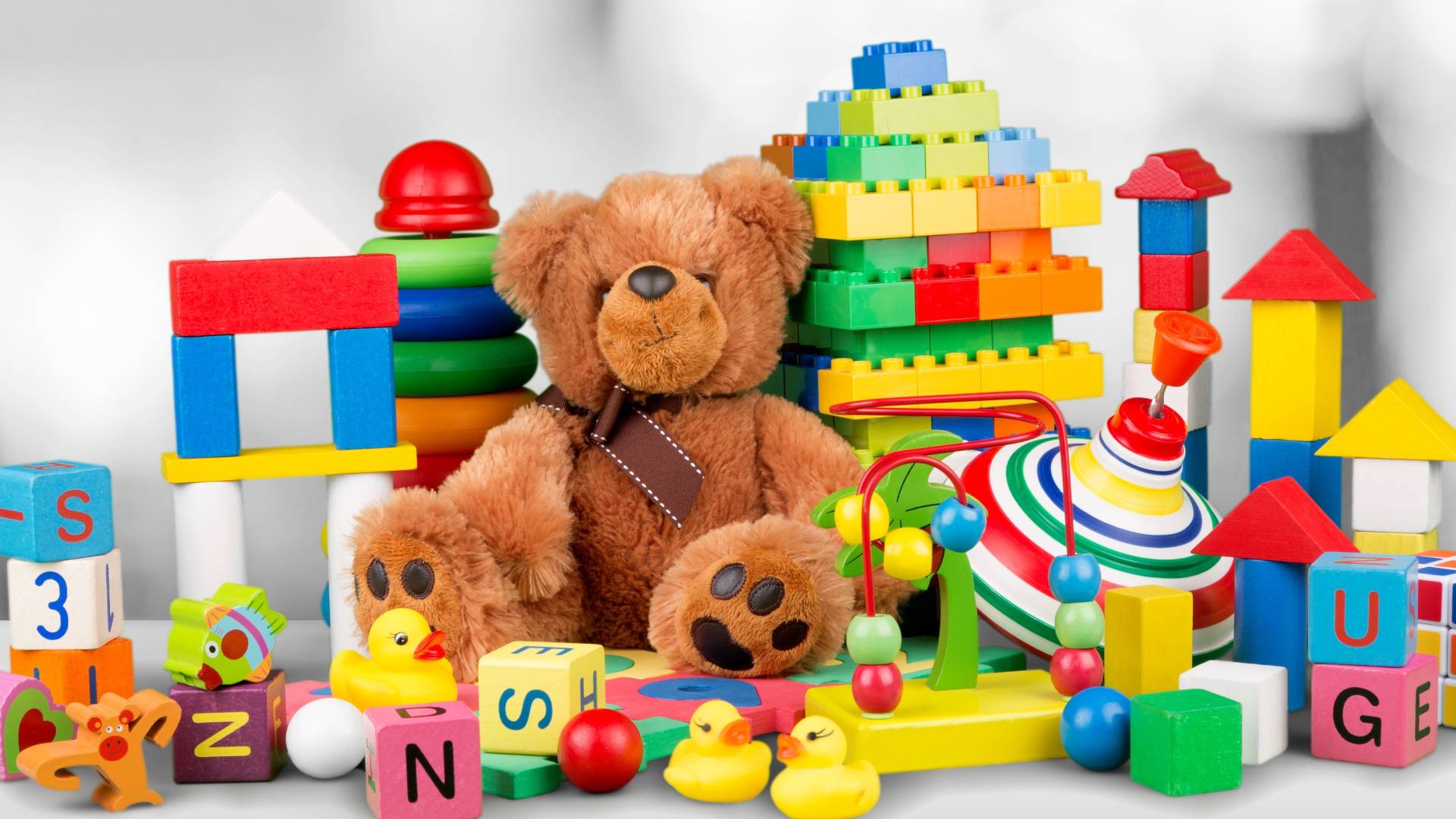 A pile of colorful toys on a plain white desk, including a stuffed bear, building blocks, alphabet blocks, and rubber ducks.
