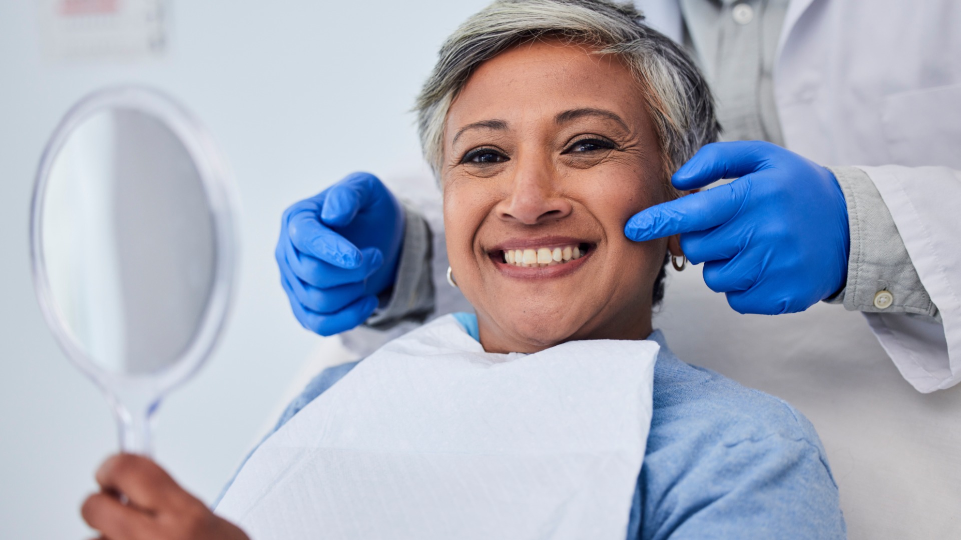 A smiling older woman sits in a dental chair holding a mirror while a dentist wearing blue gloves presses on her cheek.