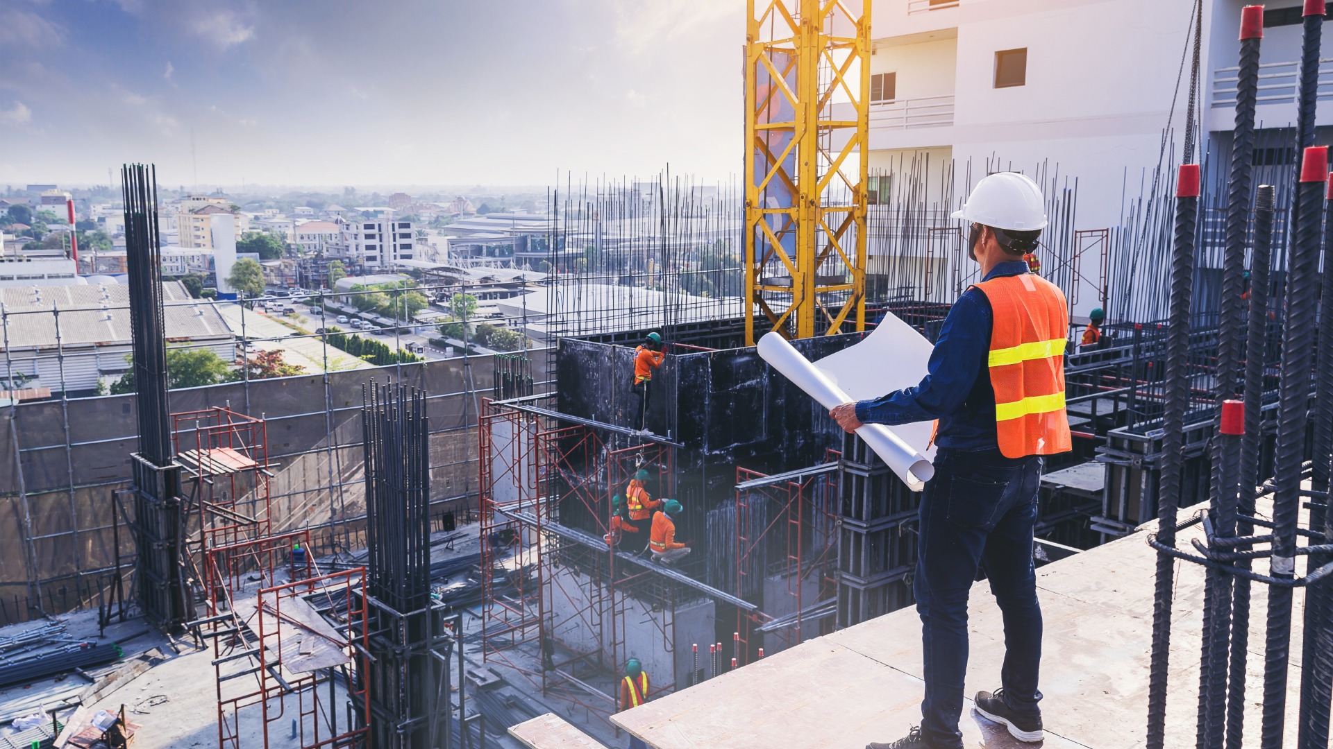 Construction engineer in an orange safety vest and a white hard hat reviewing blueprints on a high-rise building site with a crane.