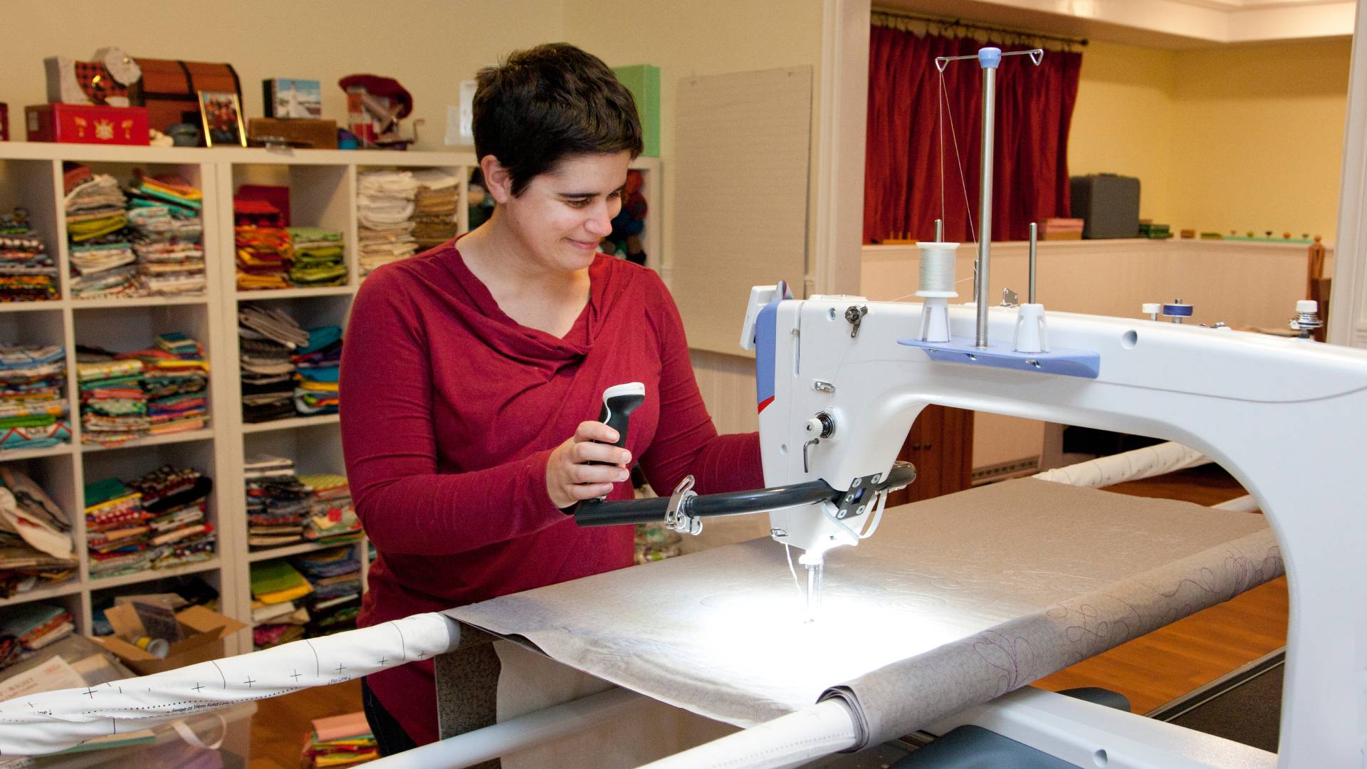 A woman in a red shirt using her embroidery machine to stitch a pattern into a large roll of decorative fabric.