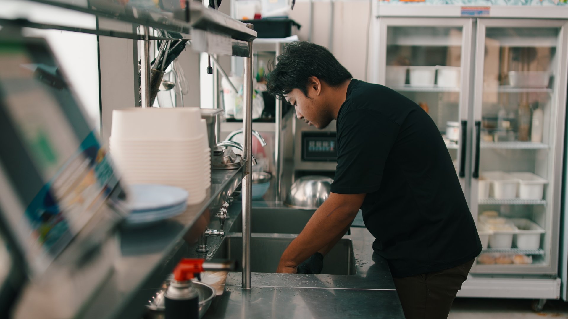 An employee wearing a black shirt reaching into a metal sink next to a POS system in a commercial kitchen.