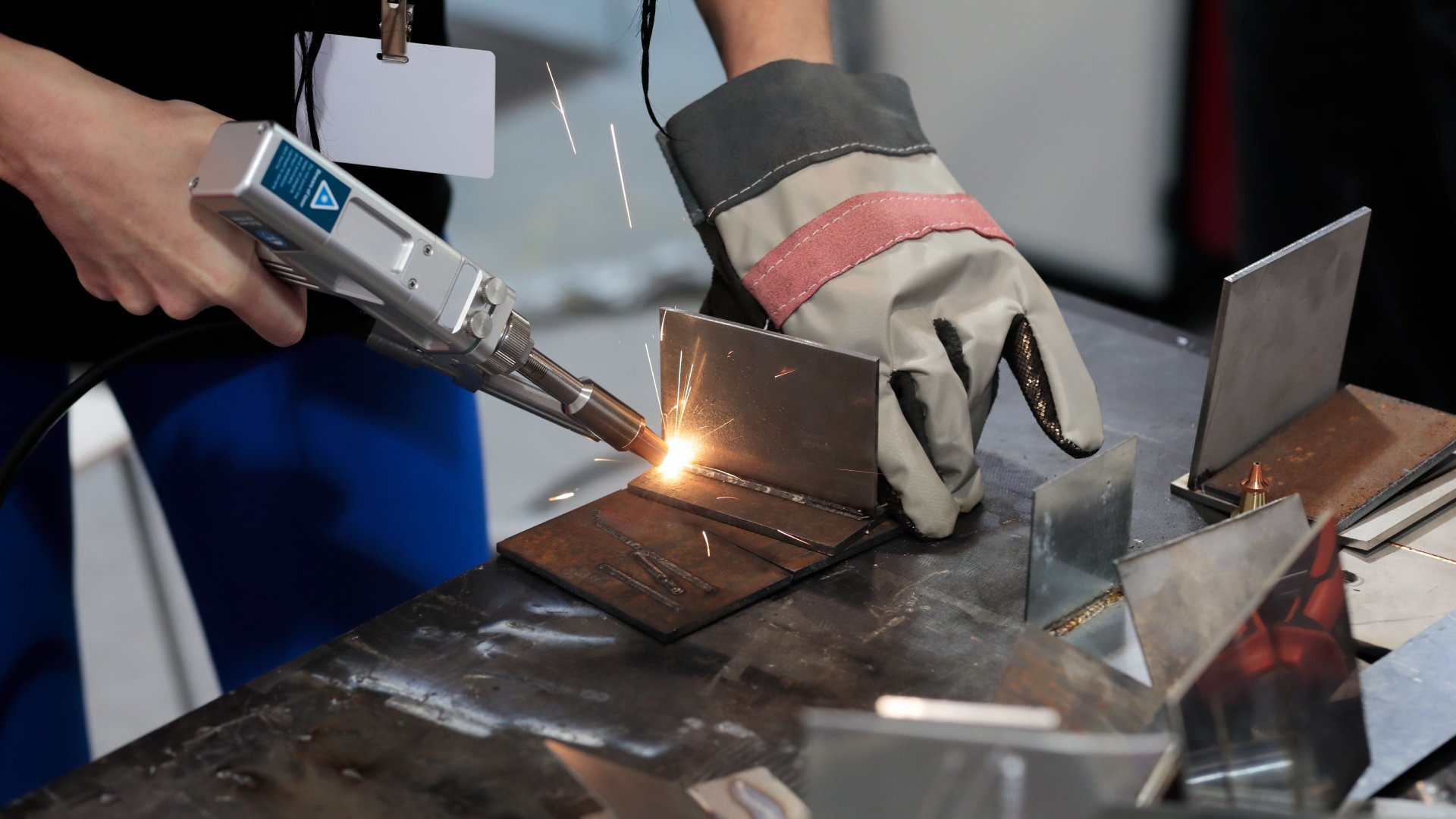 A person standing at a workstation using a portable welder to bind together pieces of metal. They're have gloves on.