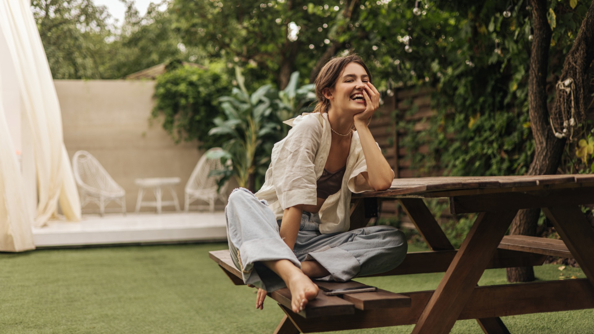 A woman sits barefoot on a wooden picnic table in a lush backyard, smiling as she rests her head in her hand.
