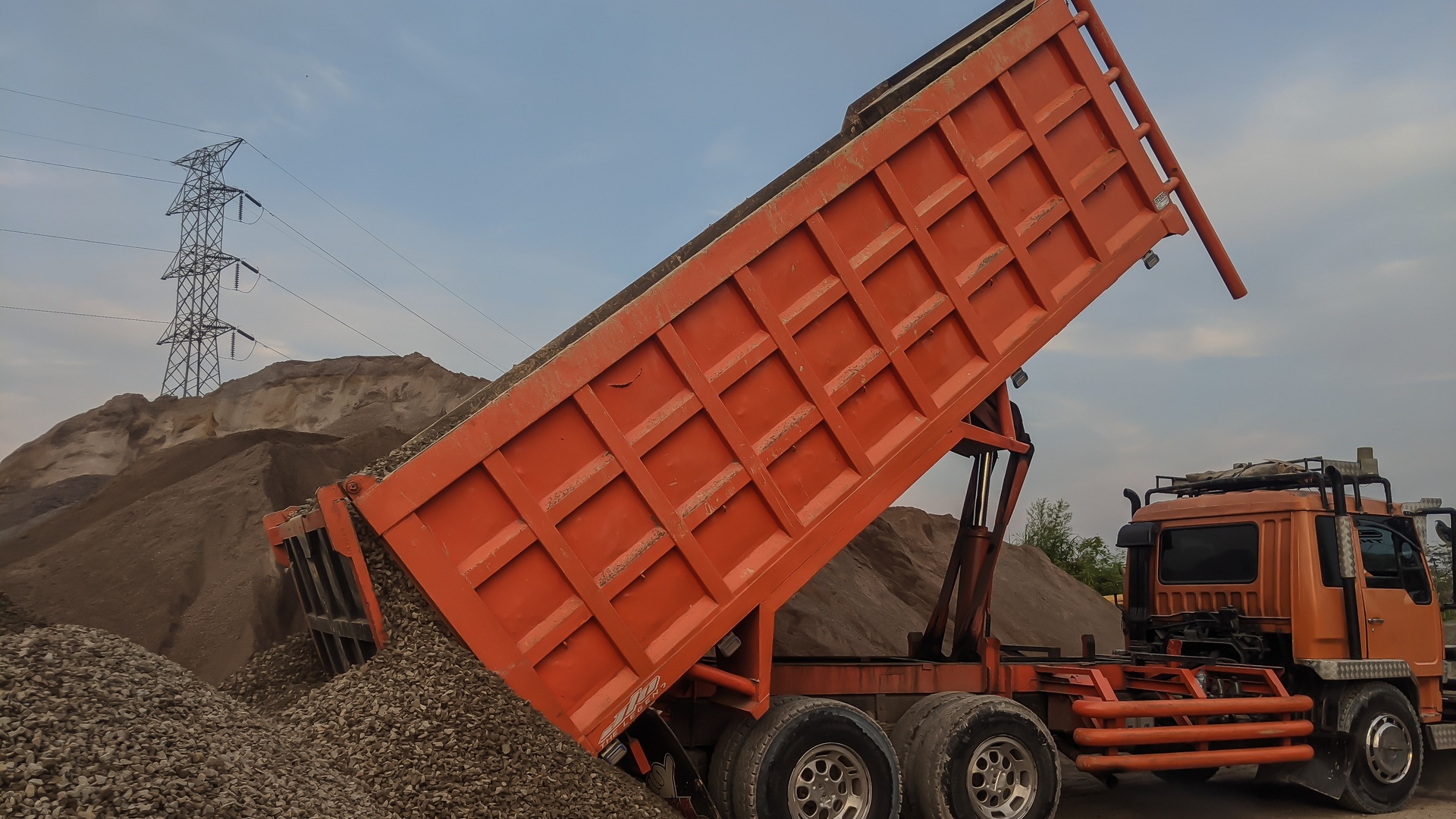 A heavy-duty, orange construction truck dumps out aggregate from the bed of its truck at a construction site.