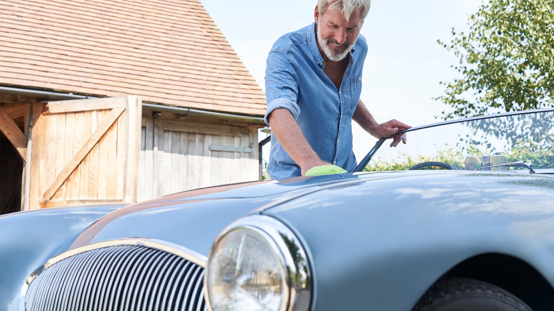 An older man with graying hair washes a light blue classic cr outdoors in front of a garage or barn building.