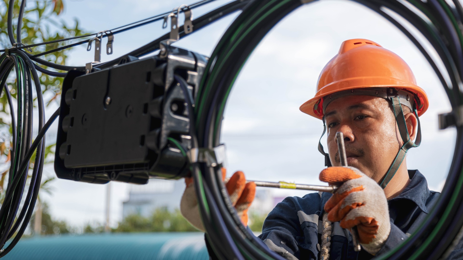A fiber optic technician outside at a fiber wire network system. They wear an orange hard hat and work on the cables.