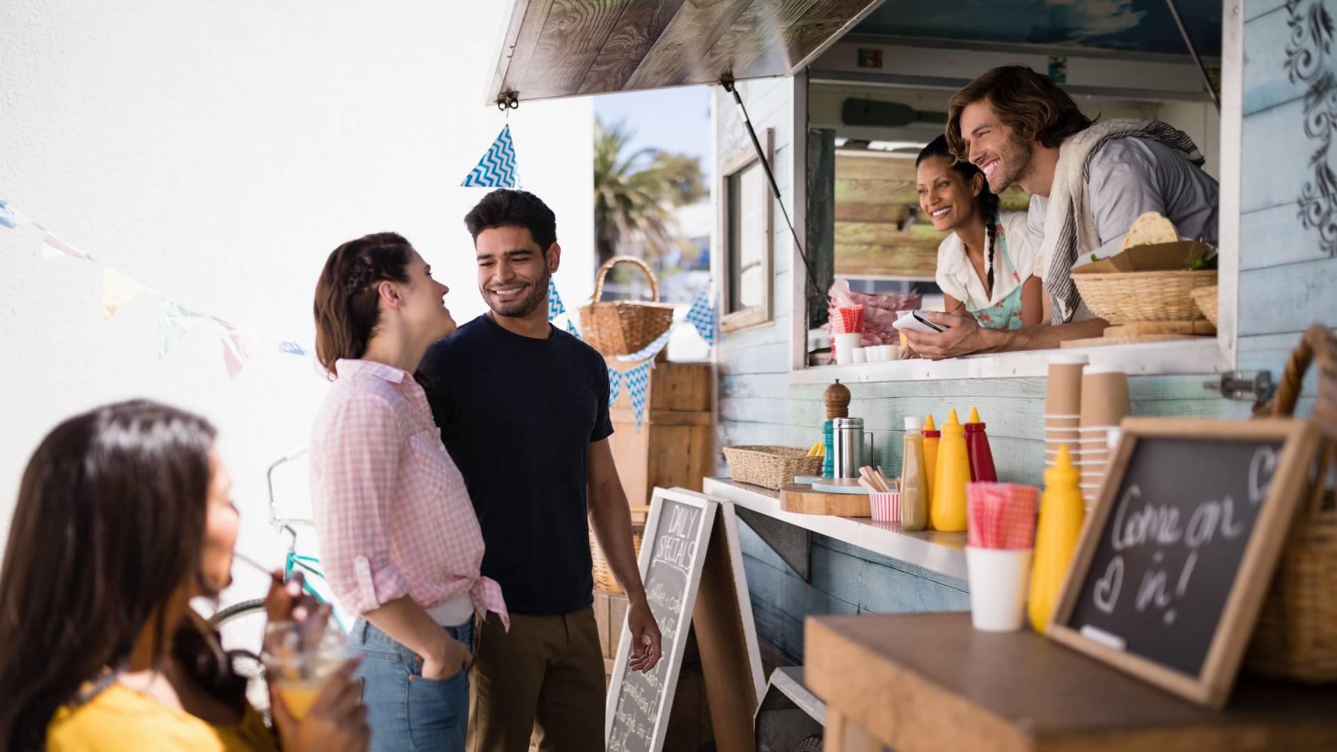 A man and woman interact with two food truck workers. Both pairs of people are smiling. On the right, a sign says, "Come on in."