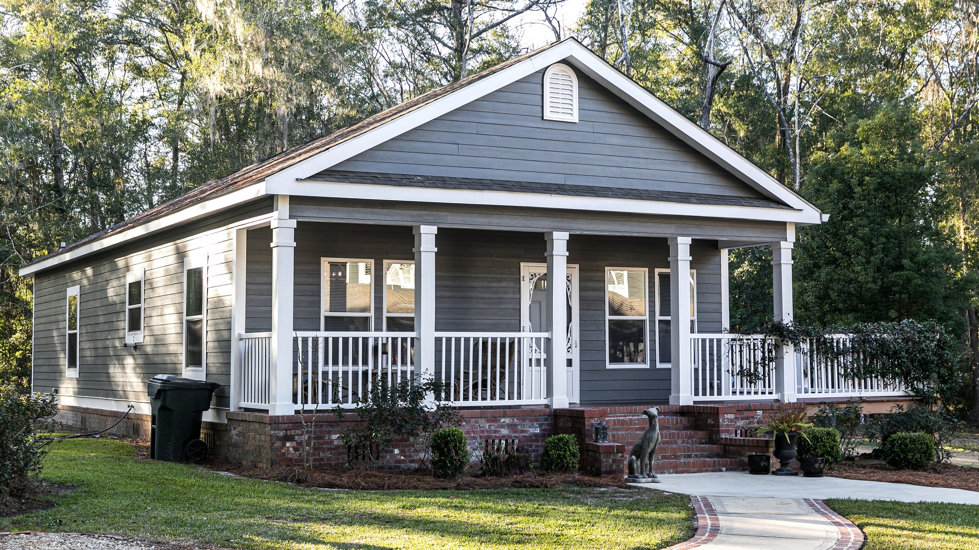 A gray mobile home with white trim, a brick foundation, and a green lawn is surrounded by trees on a sunny day.