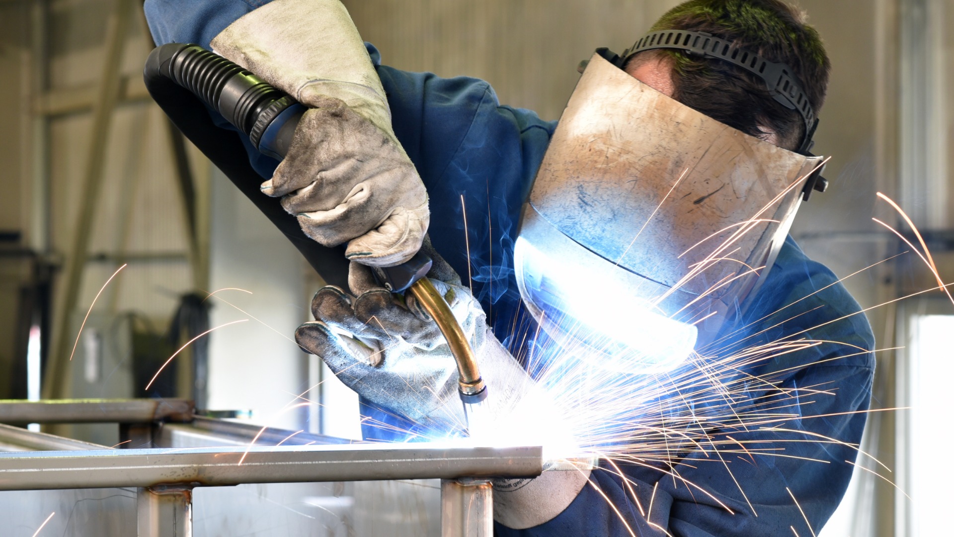 A welder wearing protective gear working on steel components with bright sparks in an industrial metal workshop.