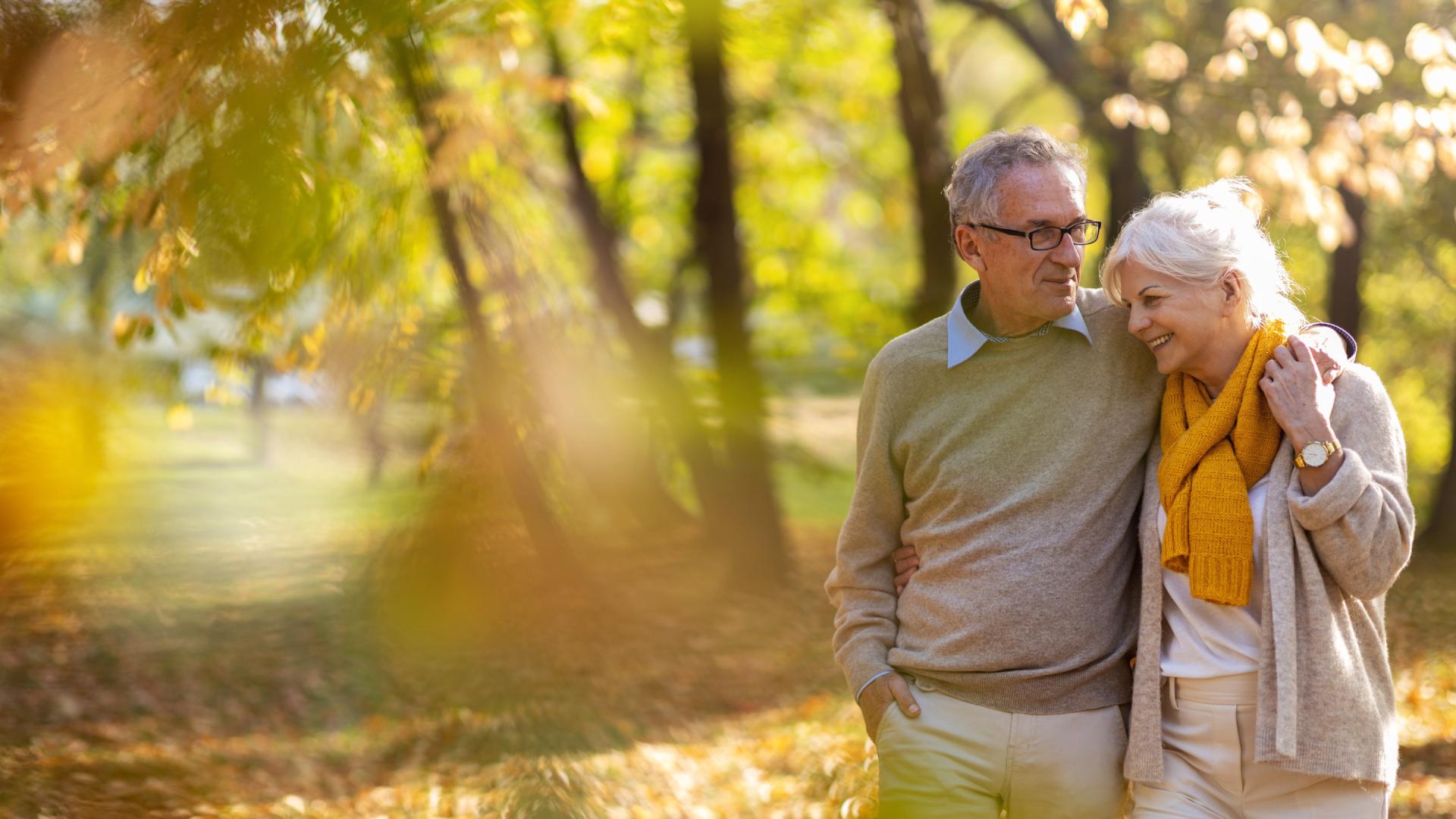 Two seniors walking through a park as leaves fall in the autumn season, wearing sweaters and scarves to stay warm.