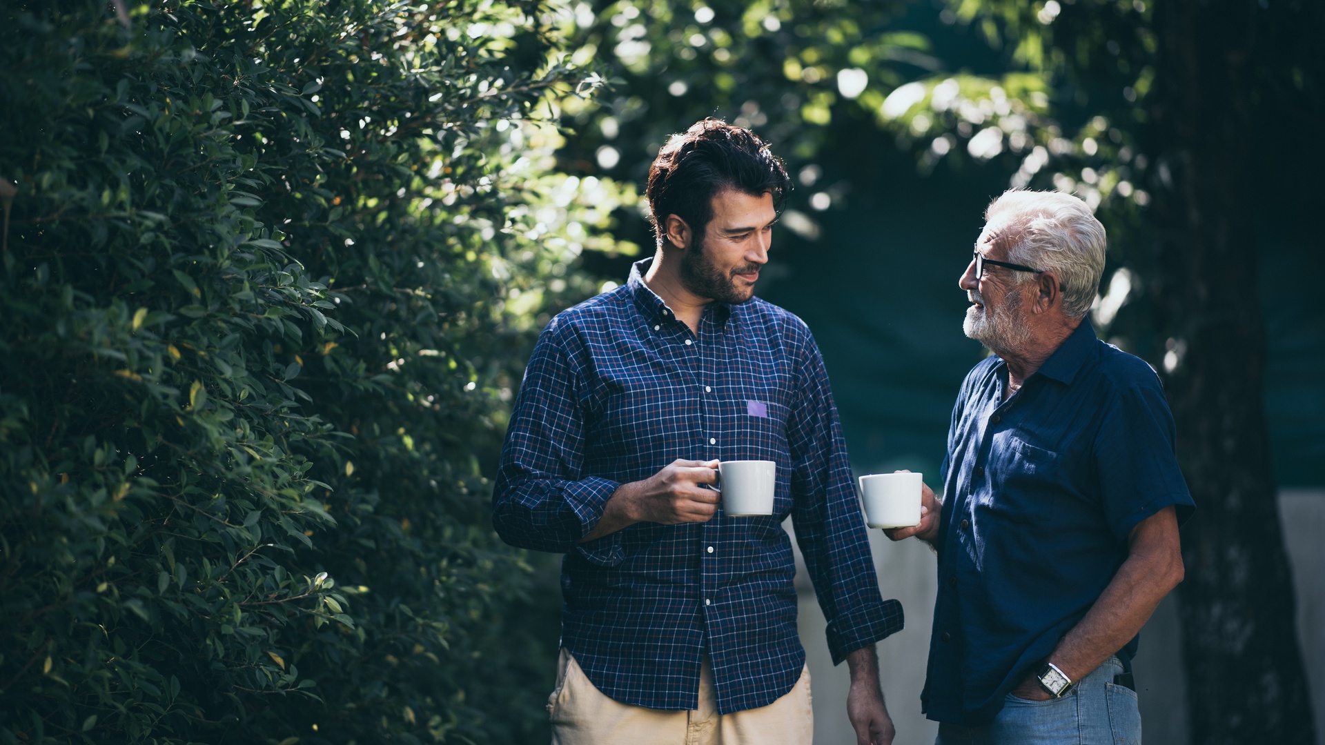 A mature father walking alongside his adult son and conversing. Both are wearing blue tops and drinking coffee.