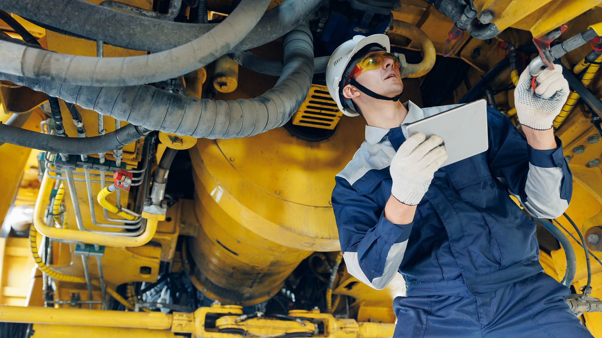 A man in a hard hat and protective glasses standing underneath a large piece of equipment. He is inspecting it.