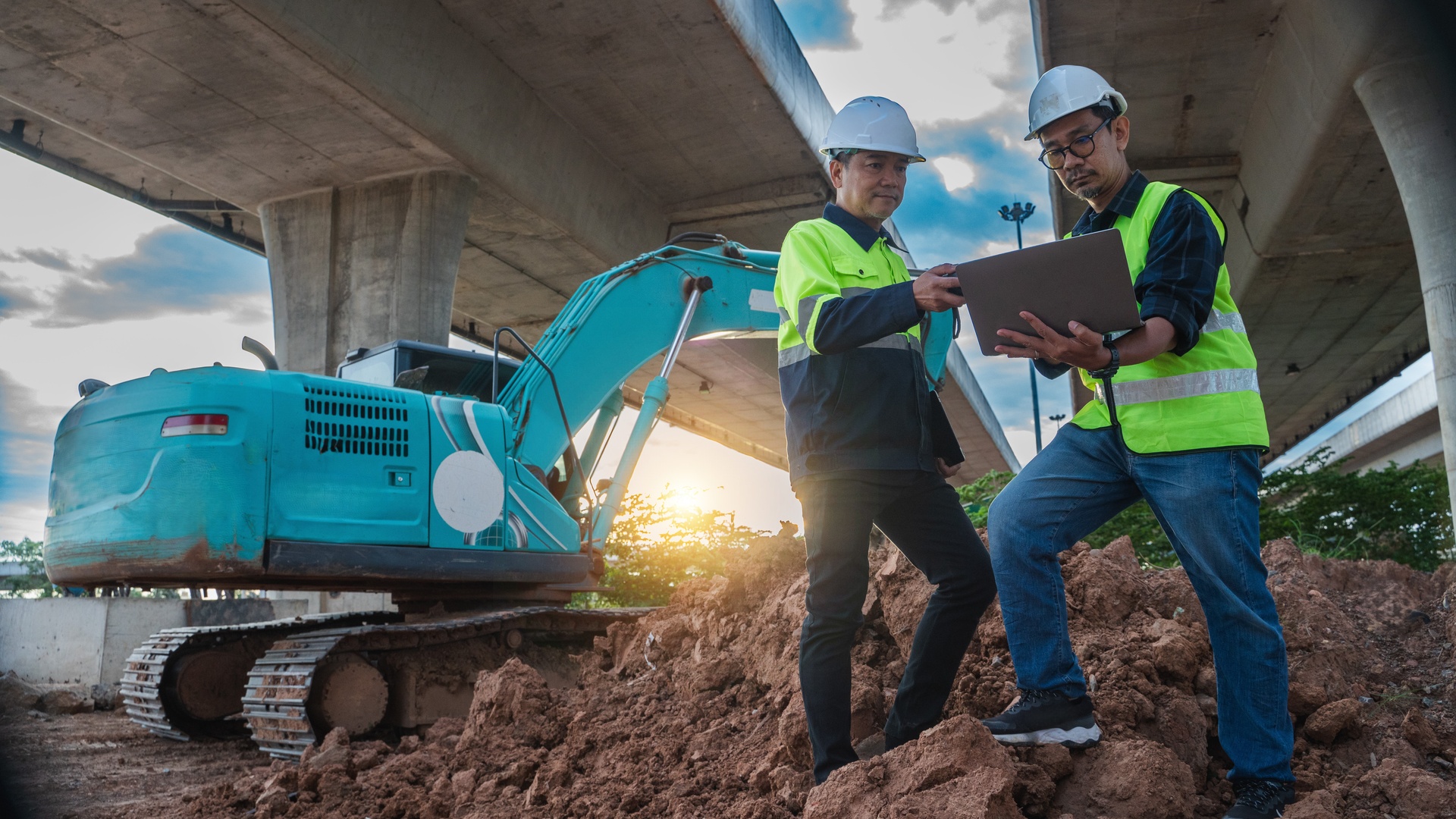 Two men in construction gear standing on a mound of dirt while looking at a laptop. There is a blue excavator behind them.