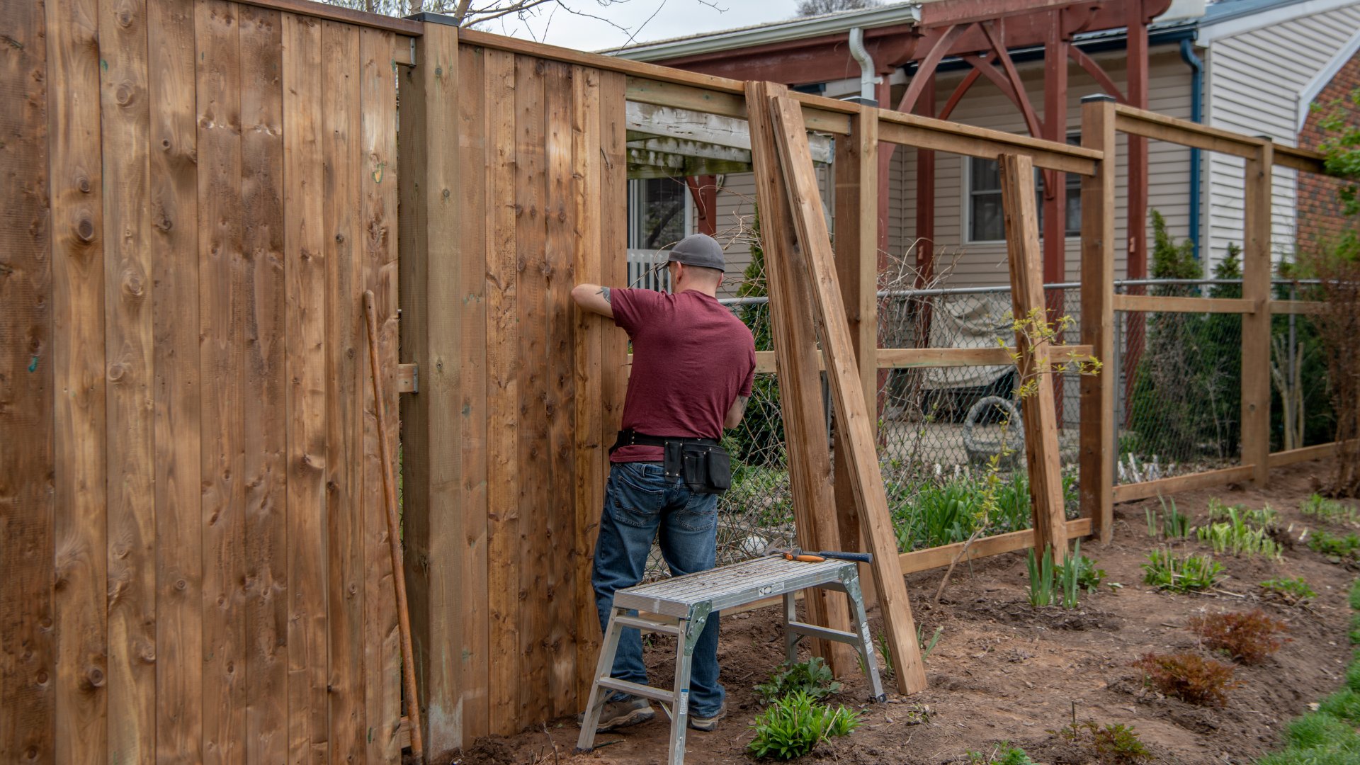A contractor wearing a gray hat and red shirt installing a tall wooden fence in a residential backyard.