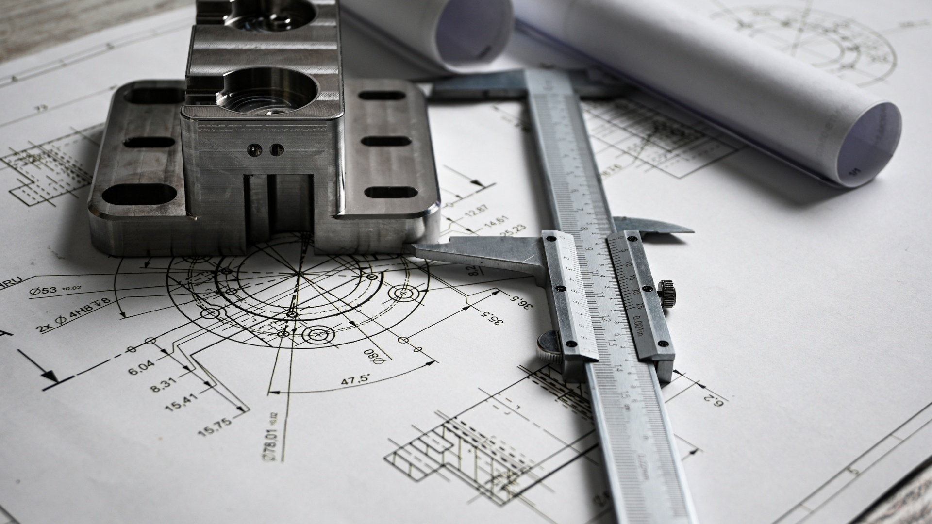 A caliper, a machined metal part, and two rolled paper sheets sit on a surface covered with a technical drawing.