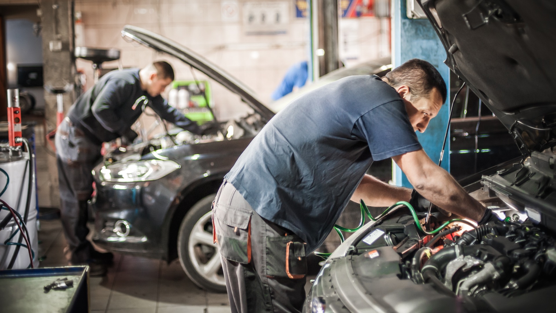 Auto mechanics under the hoods of cars in the repair shop. They each wear a mechanic's uniform and are focused.