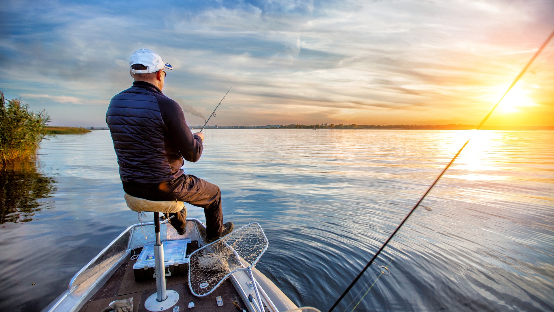 A man sits in a chair on the bow of his boat. He is holding a fishing rod and looking out at the water.