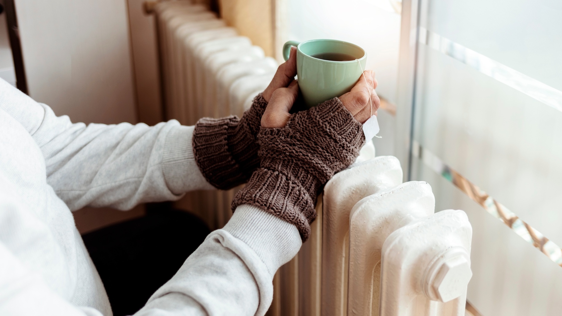 A person stands next to their old radiator wearing gloves and holding a coffee mug over the top of the radiator.