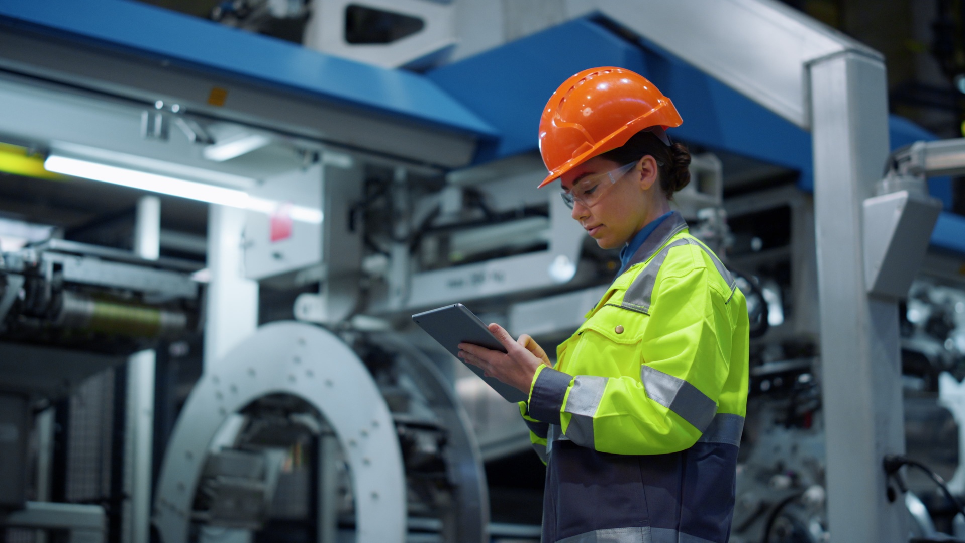 An engineer wearing a hard hat and safety goggles while holding a tablet in a large industrial facility.