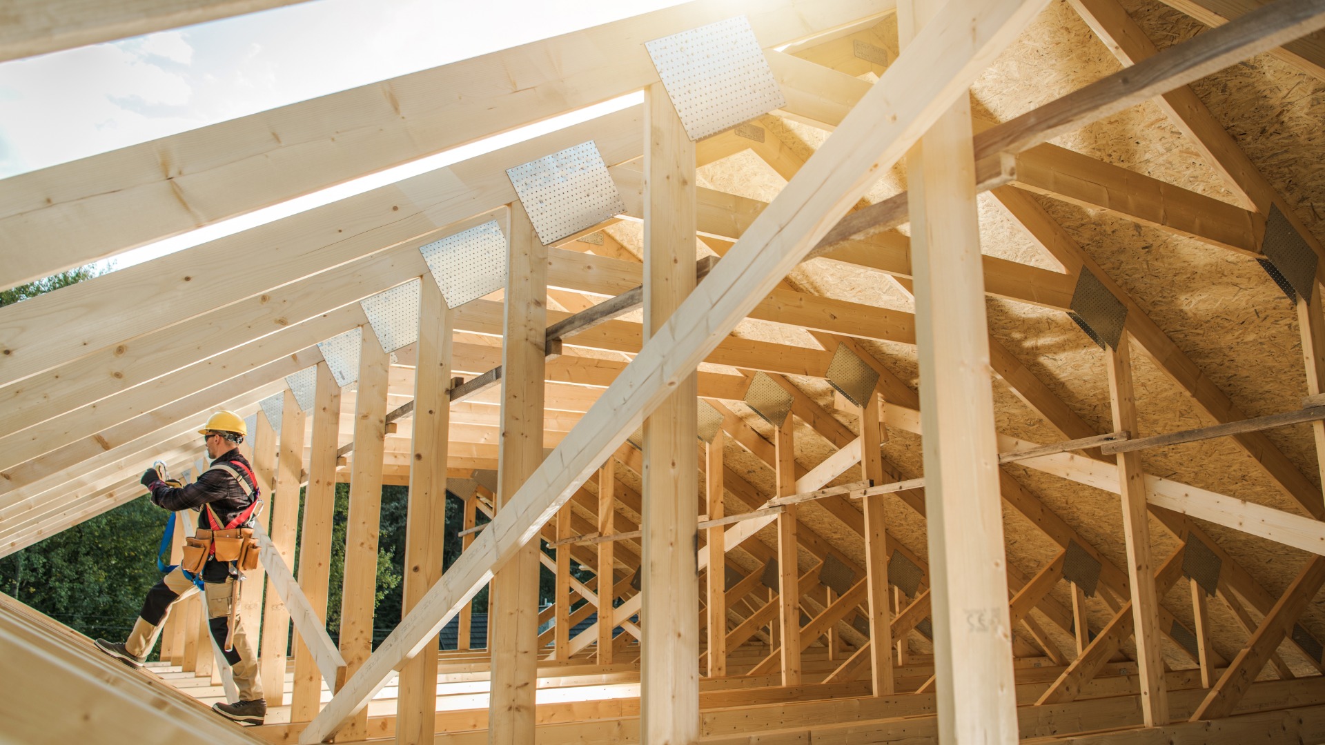 A worker securing lumber on a wooden roof frame at a residential construction site with safety gear beneath an open sky.