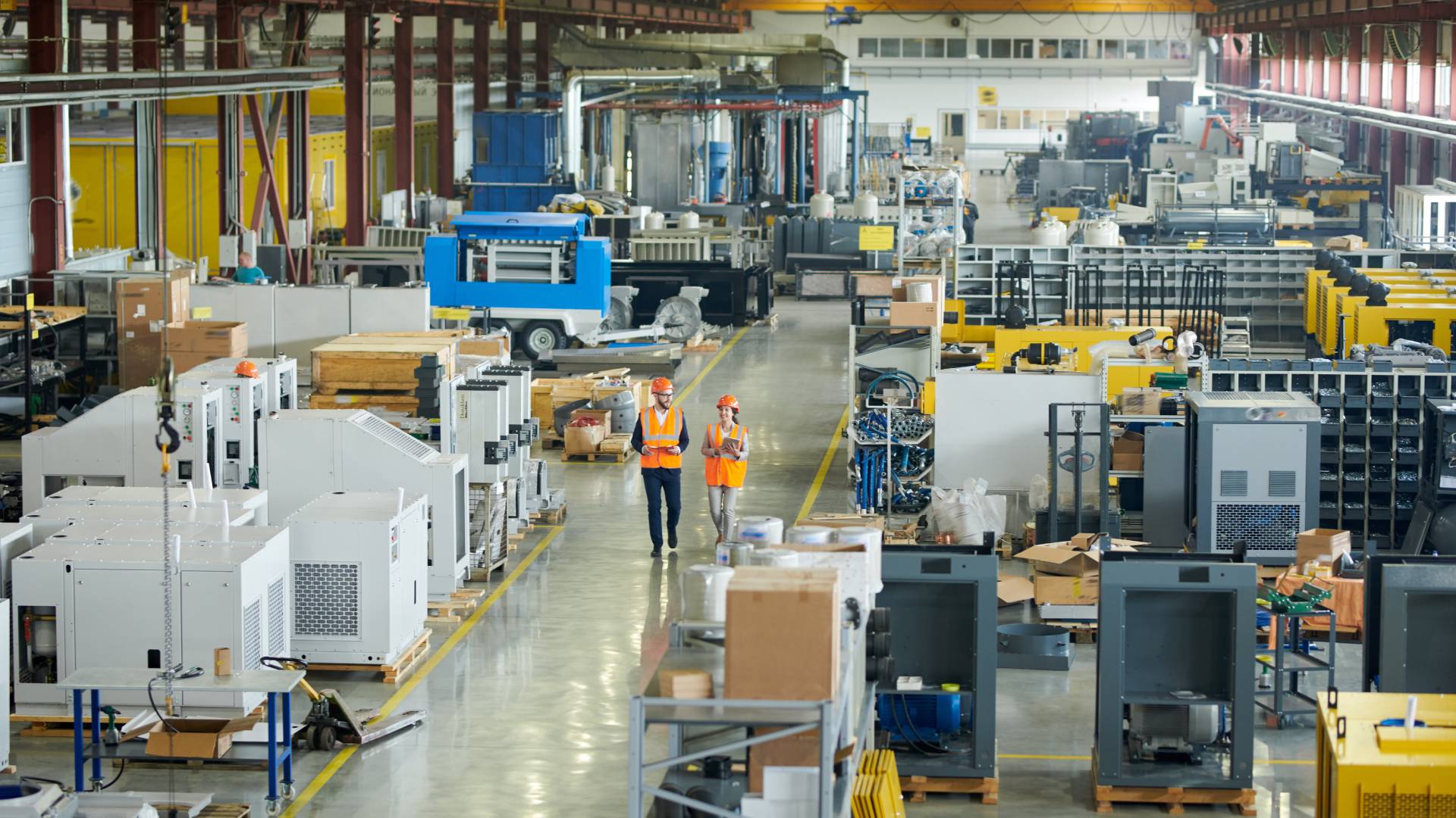 A large manufacturing facility with two employees walking down the center aisle. They're in orange safety gear.