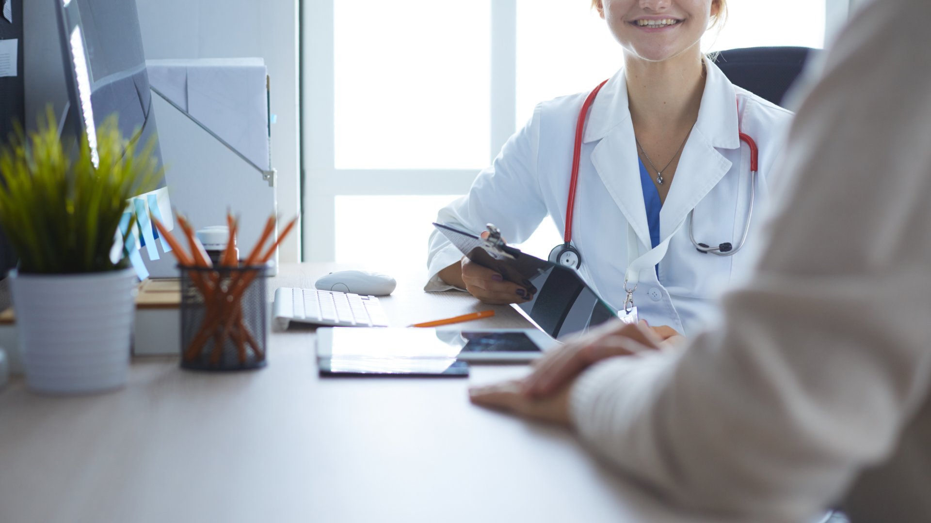 A doctor reviews notes on a clipboard while speaking with a patient across a desk in a bright medical office.
