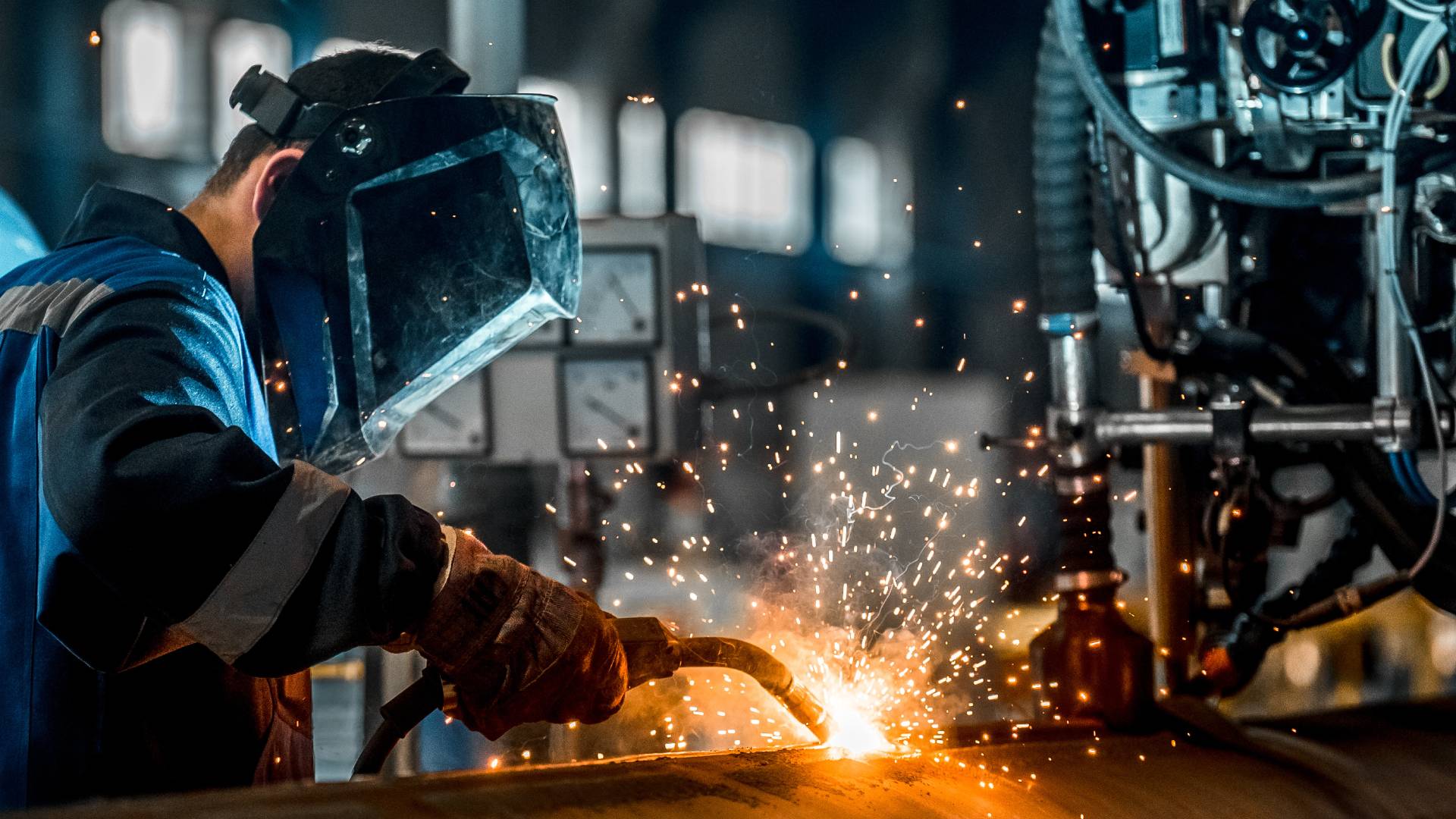 A professional welder in proper safety gear such as a helmet and gloves. They're welding a piece of metal as sparks fly.