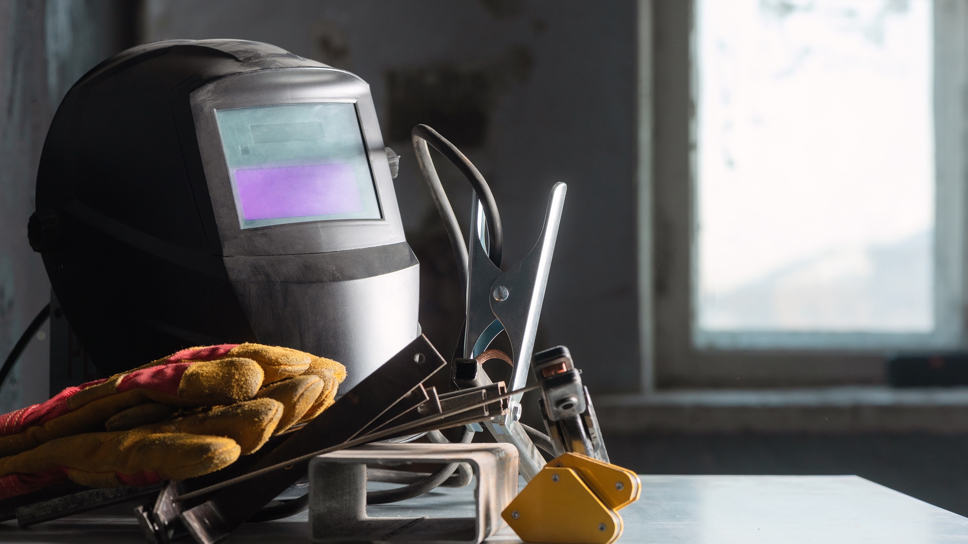 A welding helmet with a purple visor, gloves, a ground clamp, and metal pieces all lay on a silver work bench.