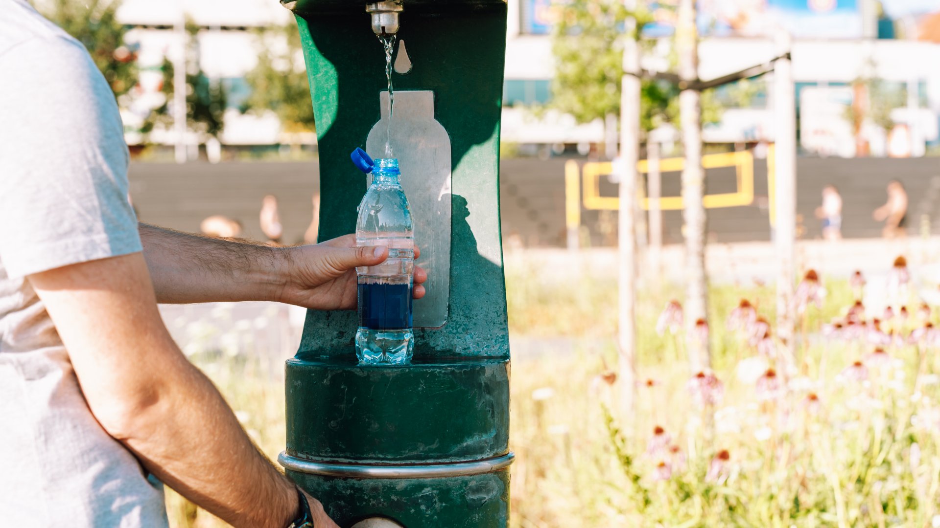 A person refilling their clear plastic water bottle at an outdoor water fountain during the daytime.