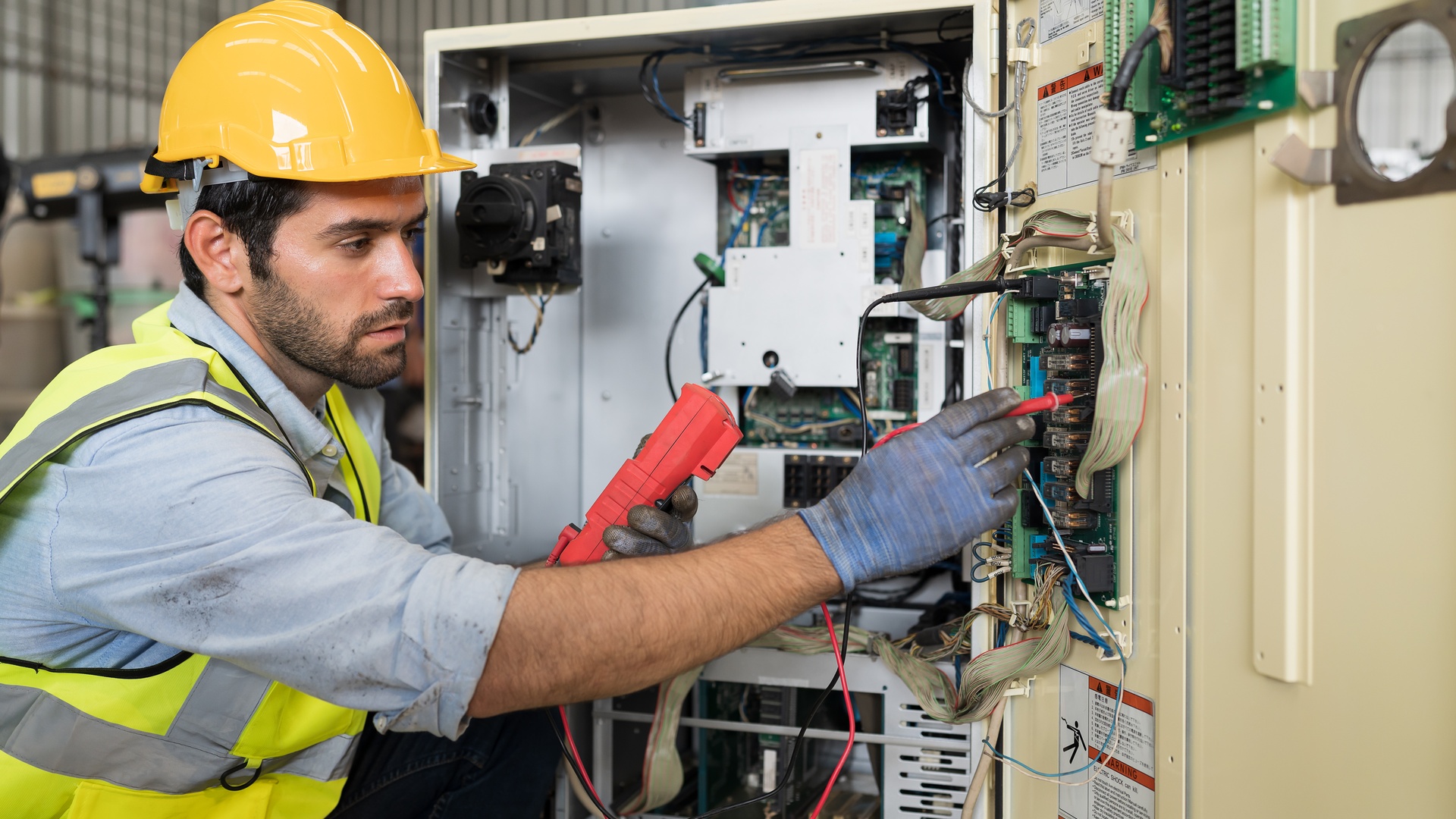 An electrician in a yellow hard hat and safety vest tests an electrical panel with a multimeter in a commercial facility.
