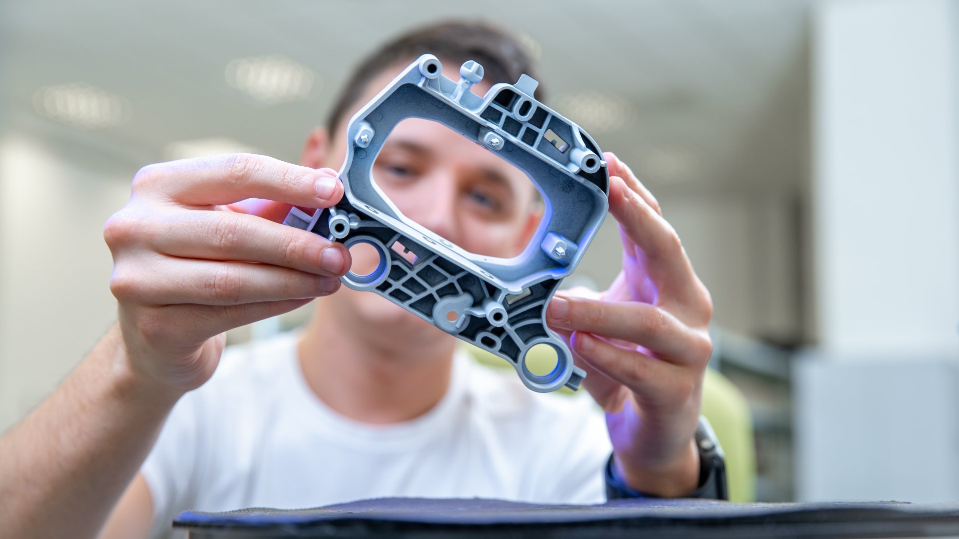 A person holds a small part of a product above a table. He is looking at the camera through the hole in the part.