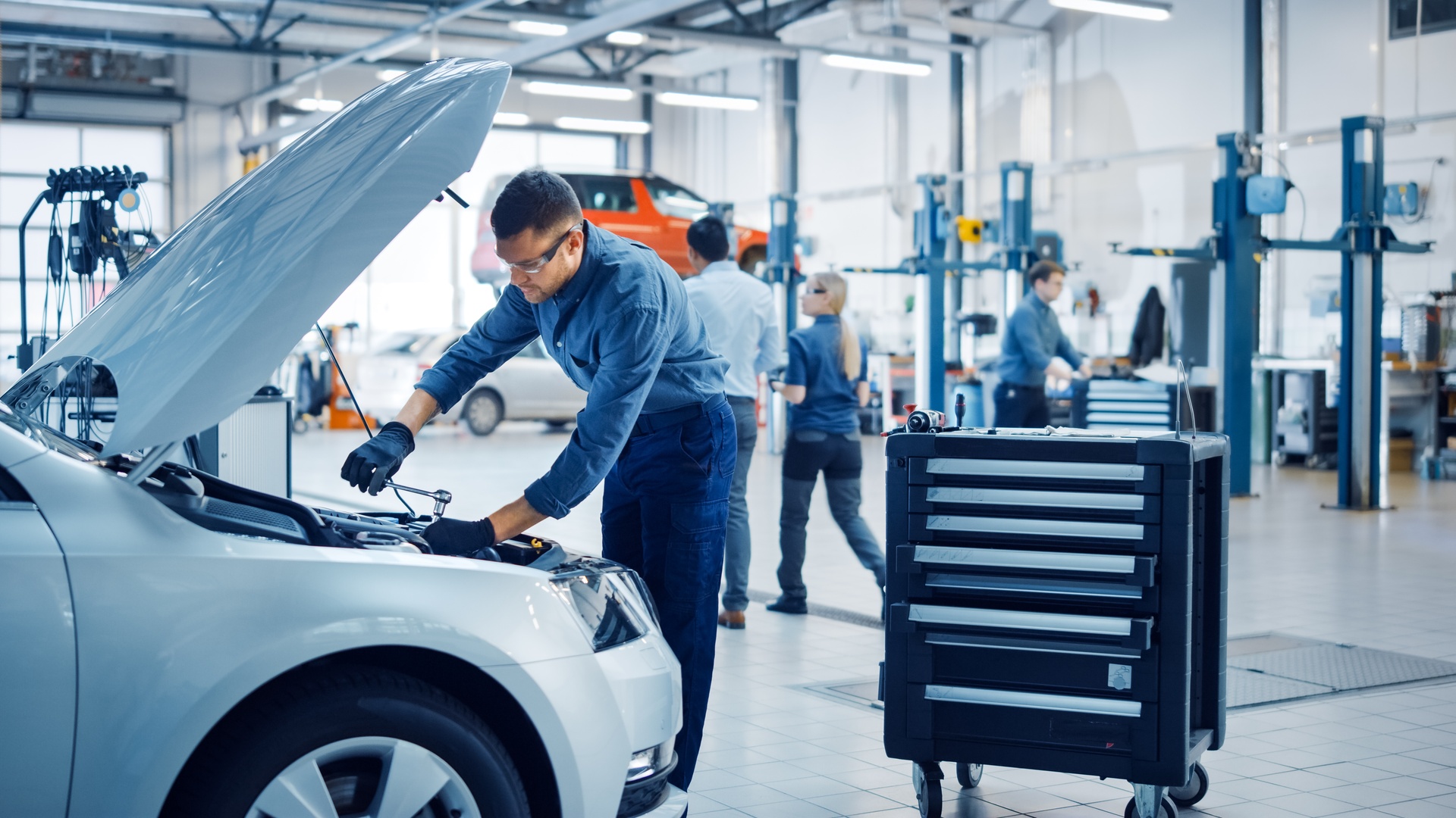 An employee working on a car engine in an auto repair shop. Three other employees walk around the shop behind them.