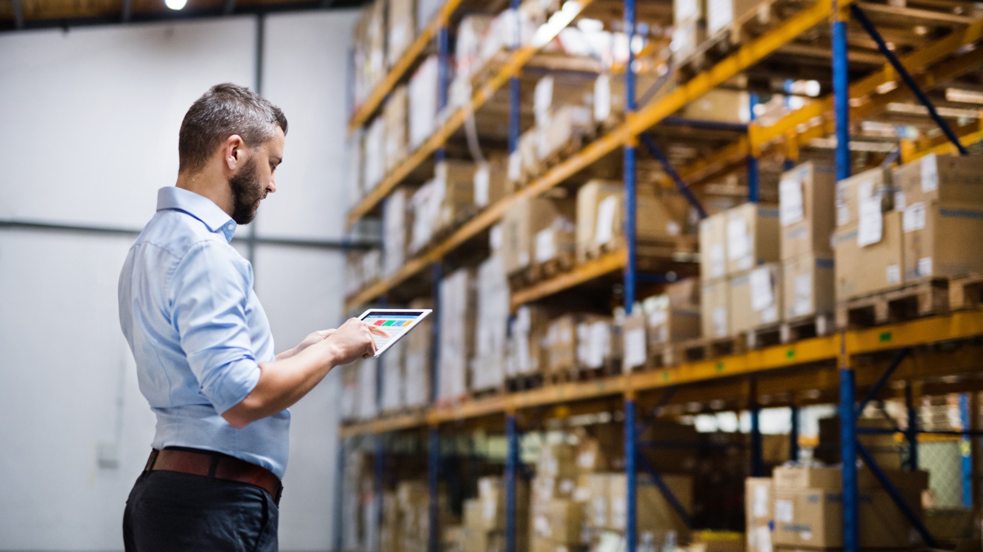An employee holding a tablet in front of a large shelving unit inside a warehouse full of cardboard boxes.