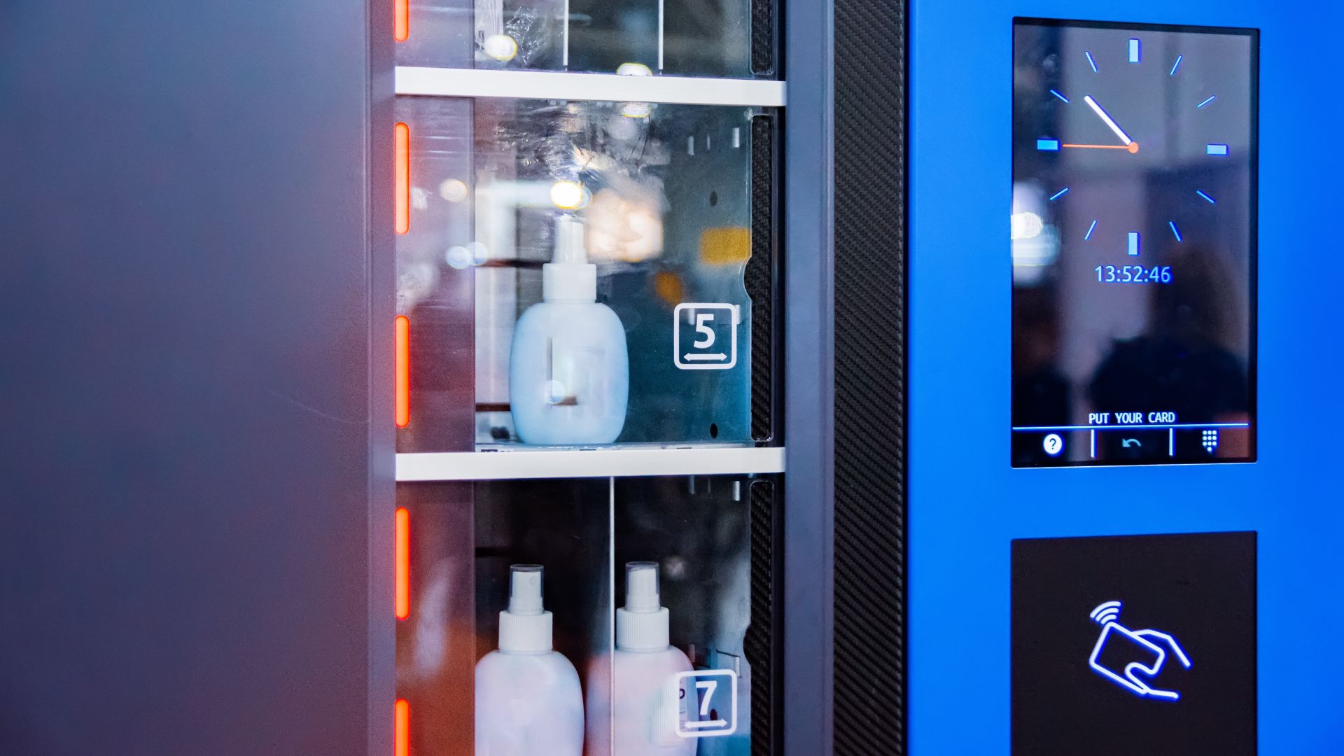 A high-tech industrial vending machine has bottles of antiseptic solution inside. There is a screen with an analog clock.