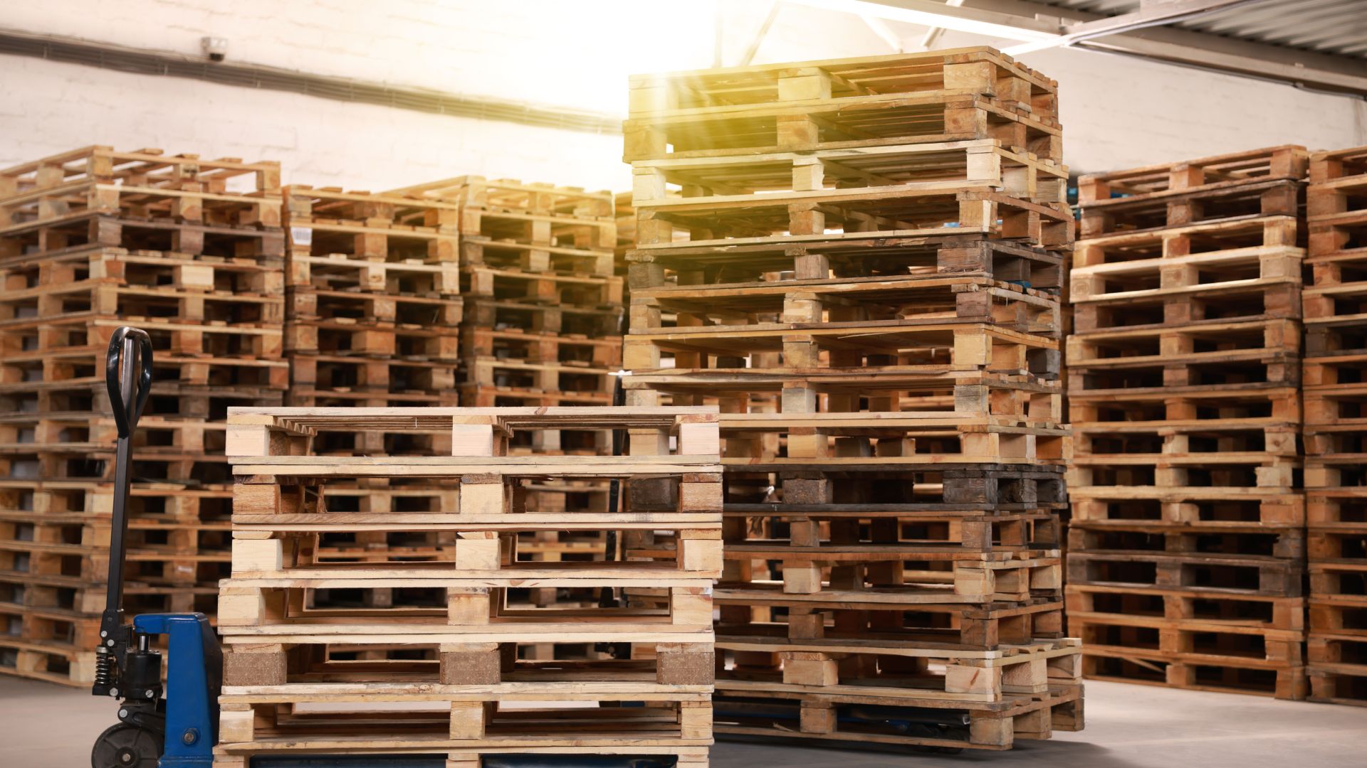 An inventory of wooden pallets stacked high in a warehousing facility. A smaller stack is on a forklift.