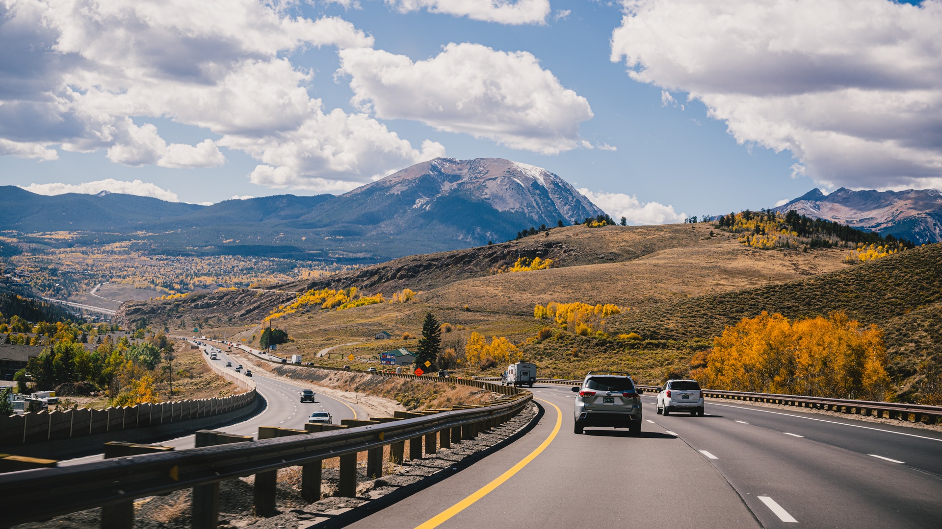 Cars drive on a curving mountain highway under a blue sky with clouds, with golden fall trees across the valley.