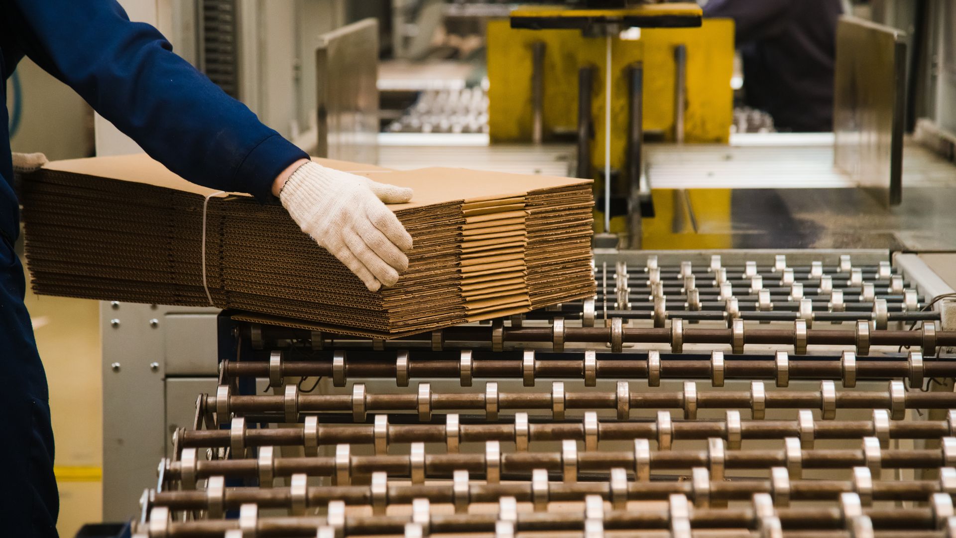 A shipping facility worker wearing safety gloves pulling a stack of corrugated cardboard boxes off the conveyor belt.