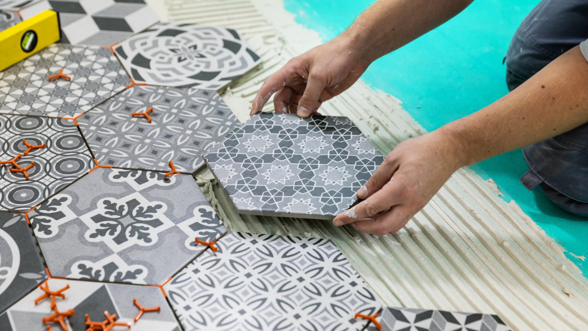 A man kneels down and lays different patterned, white and gray hexagon cement tiles on the floor of his home.