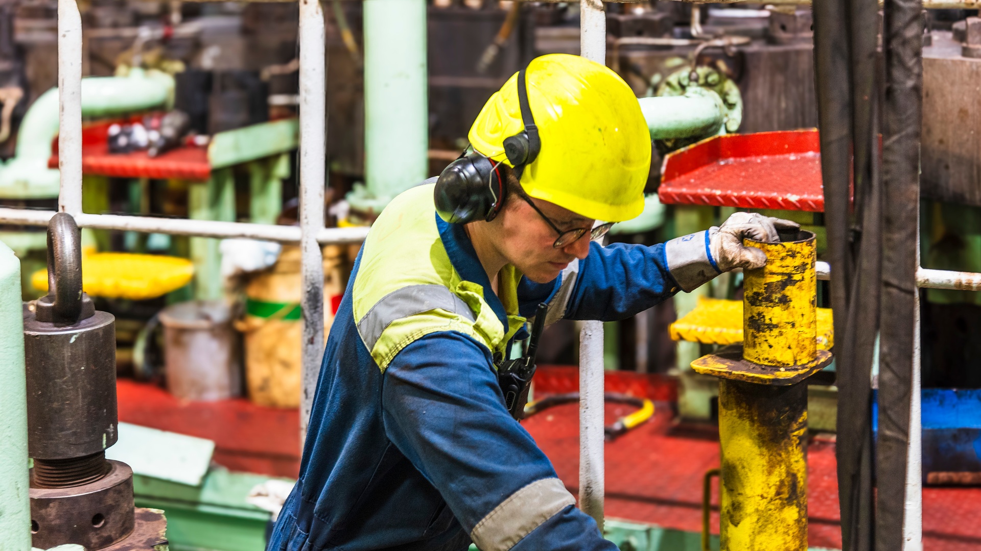 A close-up of a person wearing a hard hat and protective hearing equipment while holding a metal pipe in an engine room.